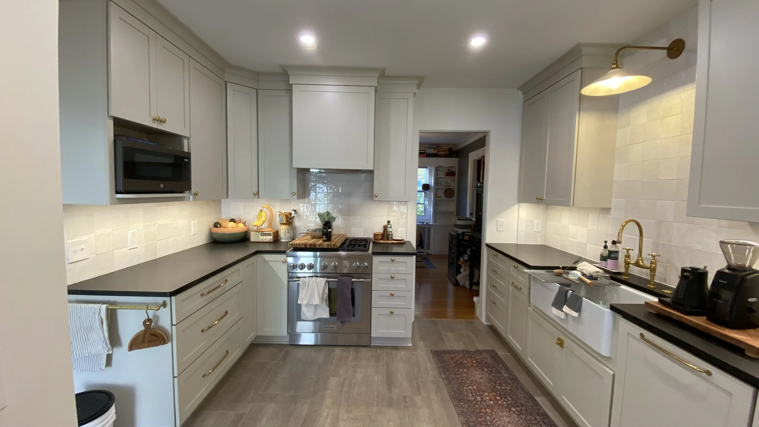 A kitchen with beige cabinets and black countertops, stainless steel oven and microwave, and small appliances on the counter. There are fruit bowls, chopping boards, and dish soap near the sink, with a doorway leading to another room. Recessed lighti