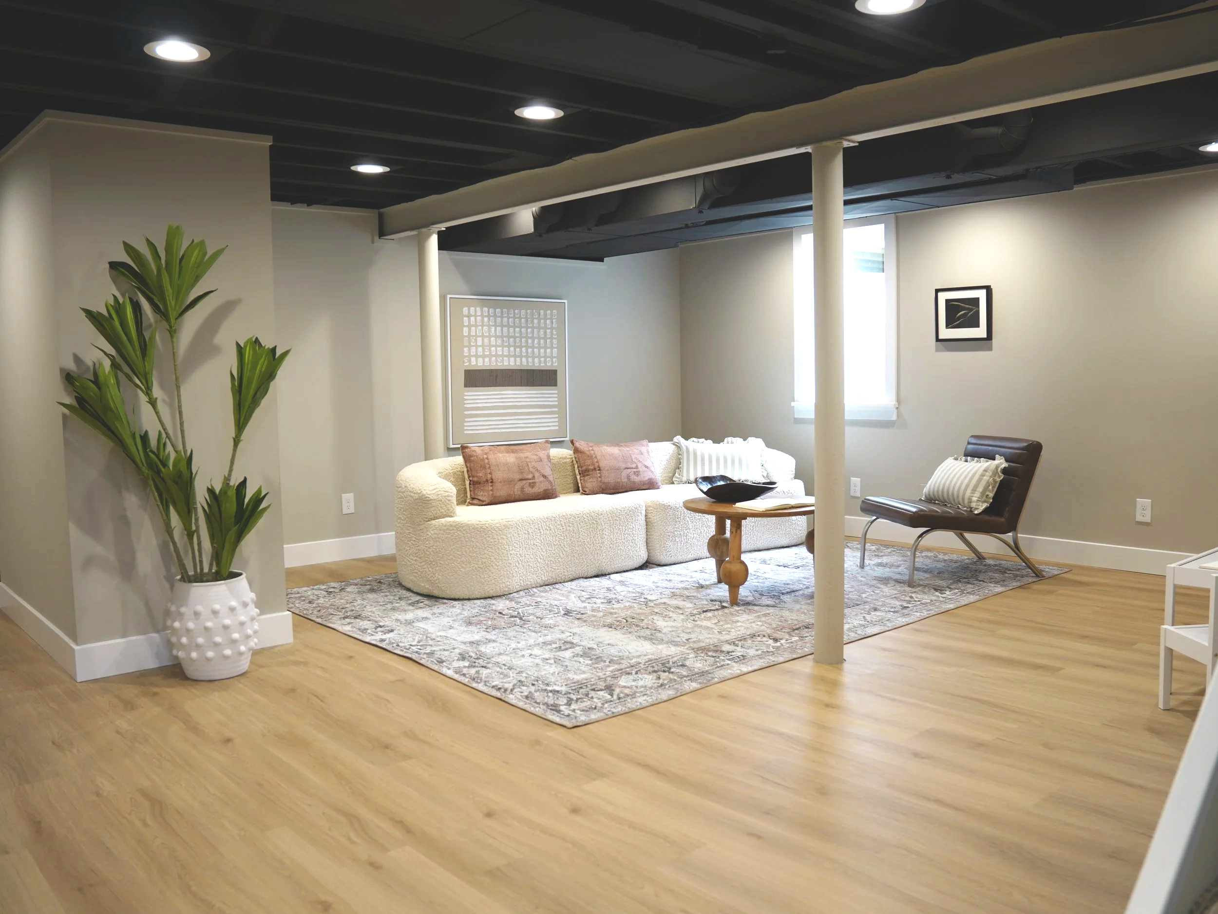 A modern living room with a beige sofa, a wooden coffee table, a black armchair, framed wall art, a large green plant in a white pot, and a patterned area rug on hardwood flooring.