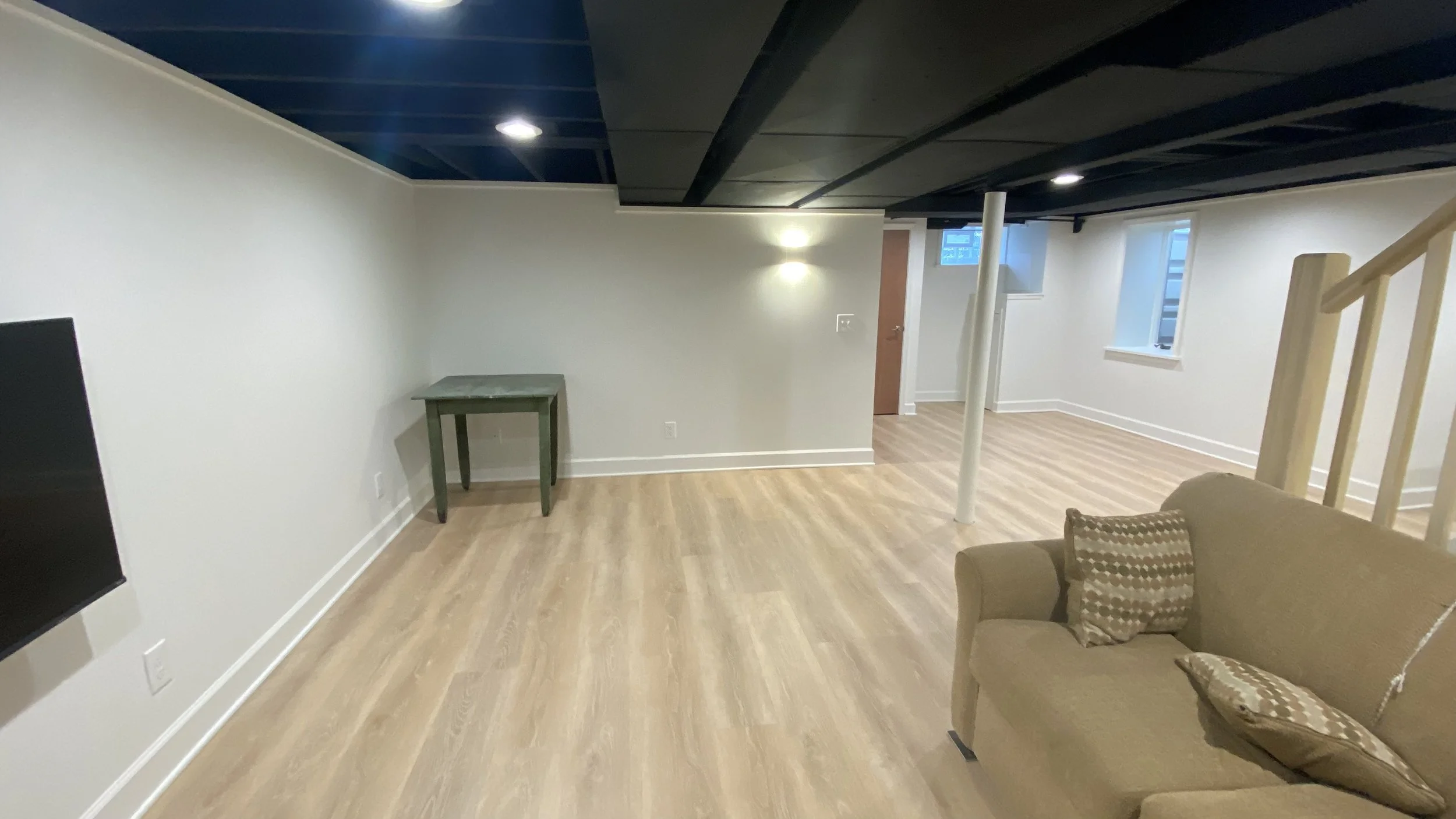 A spacious basement room with light wood flooring, white walls, and a black ceiling. There are windows allowing natural light, a small green table, an armchair with patterned cushions, and a partial view of a staircase.