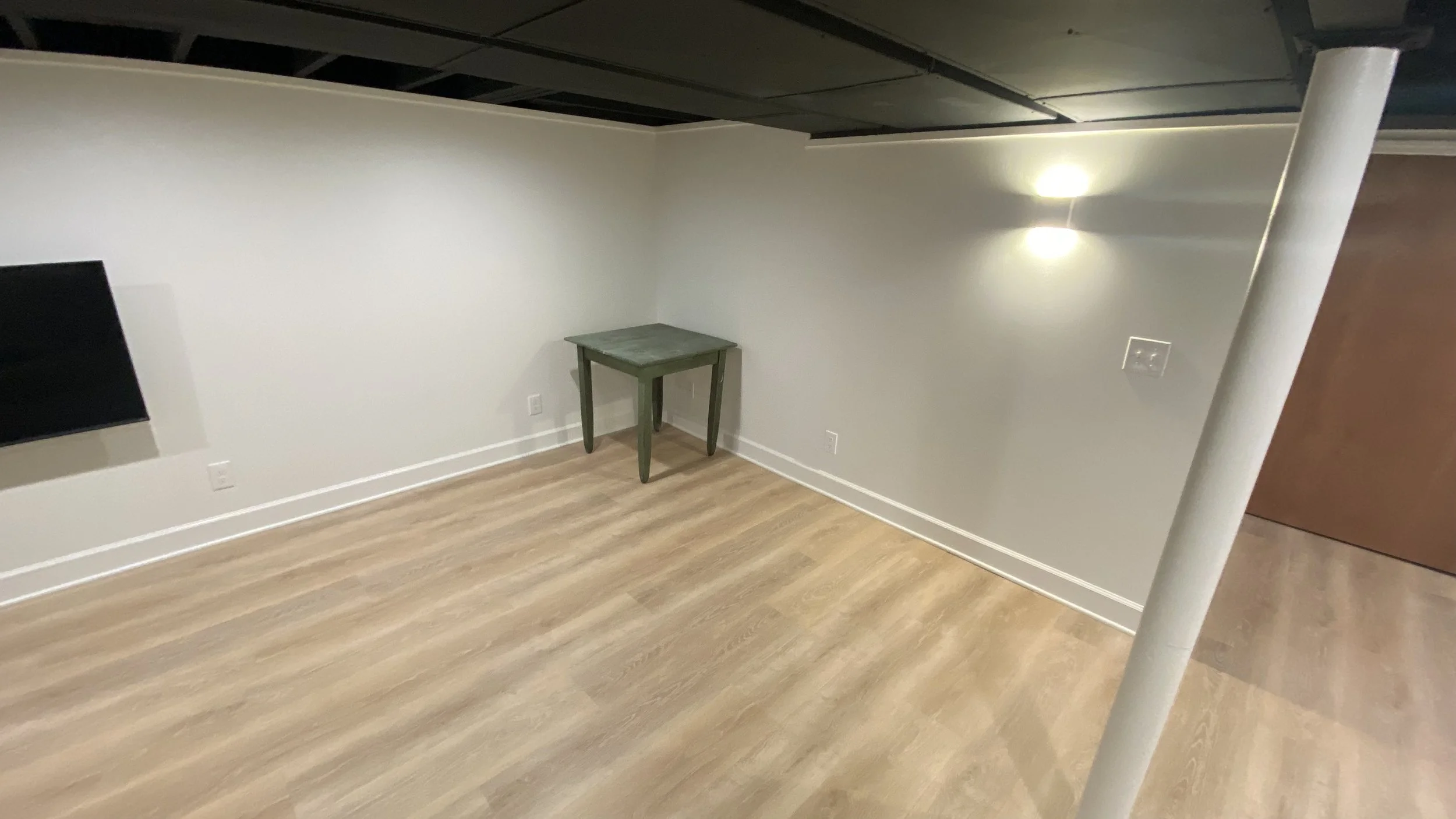 Empty room with white walls, wood flooring, a small green table, and a wall-mounted TV. There are two light fixtures on the right wall and visible ceiling beams.