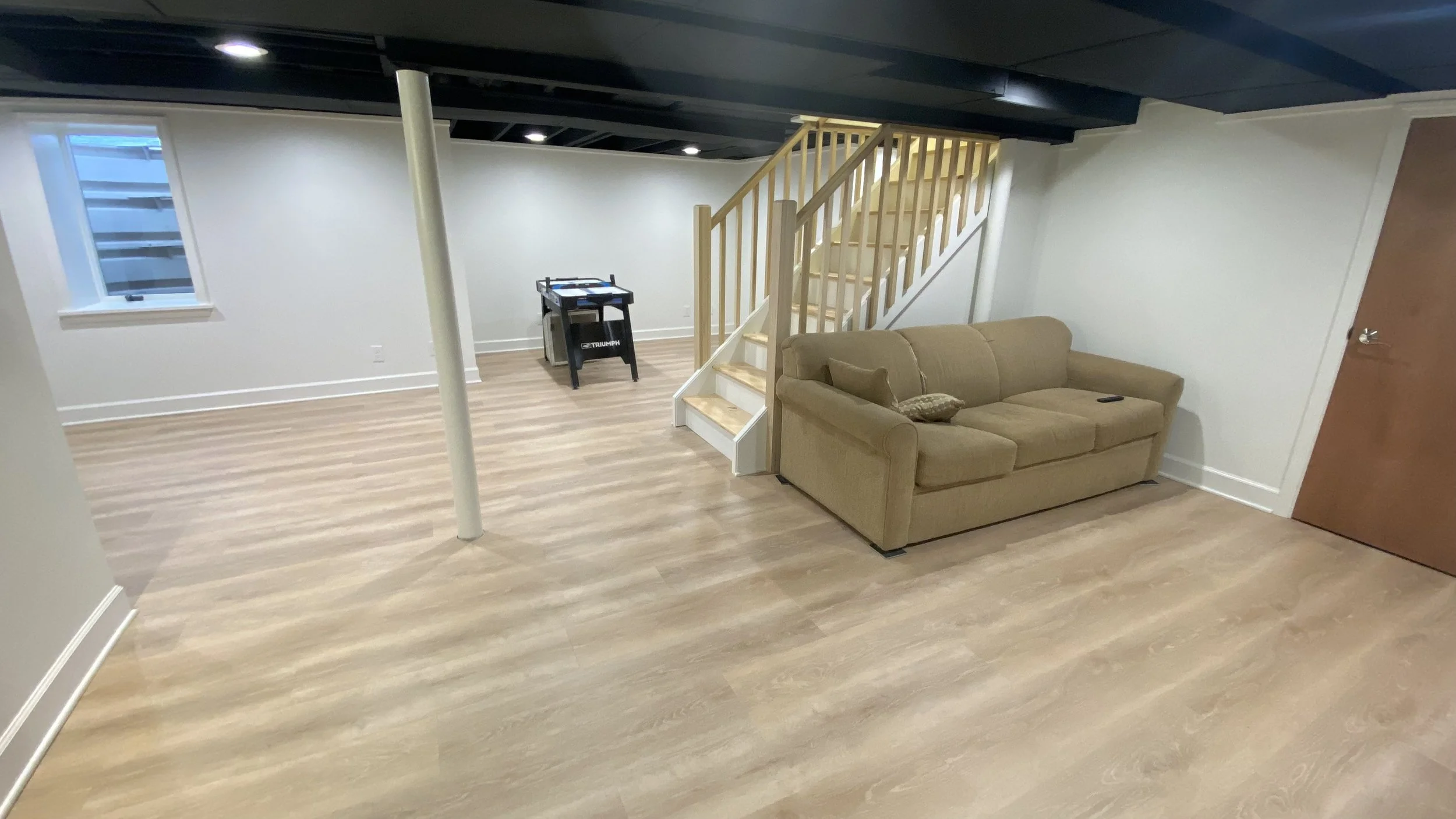 Living room with beige couch, wooden staircase, light wood laminate flooring, white walls, window, and a foosball table.