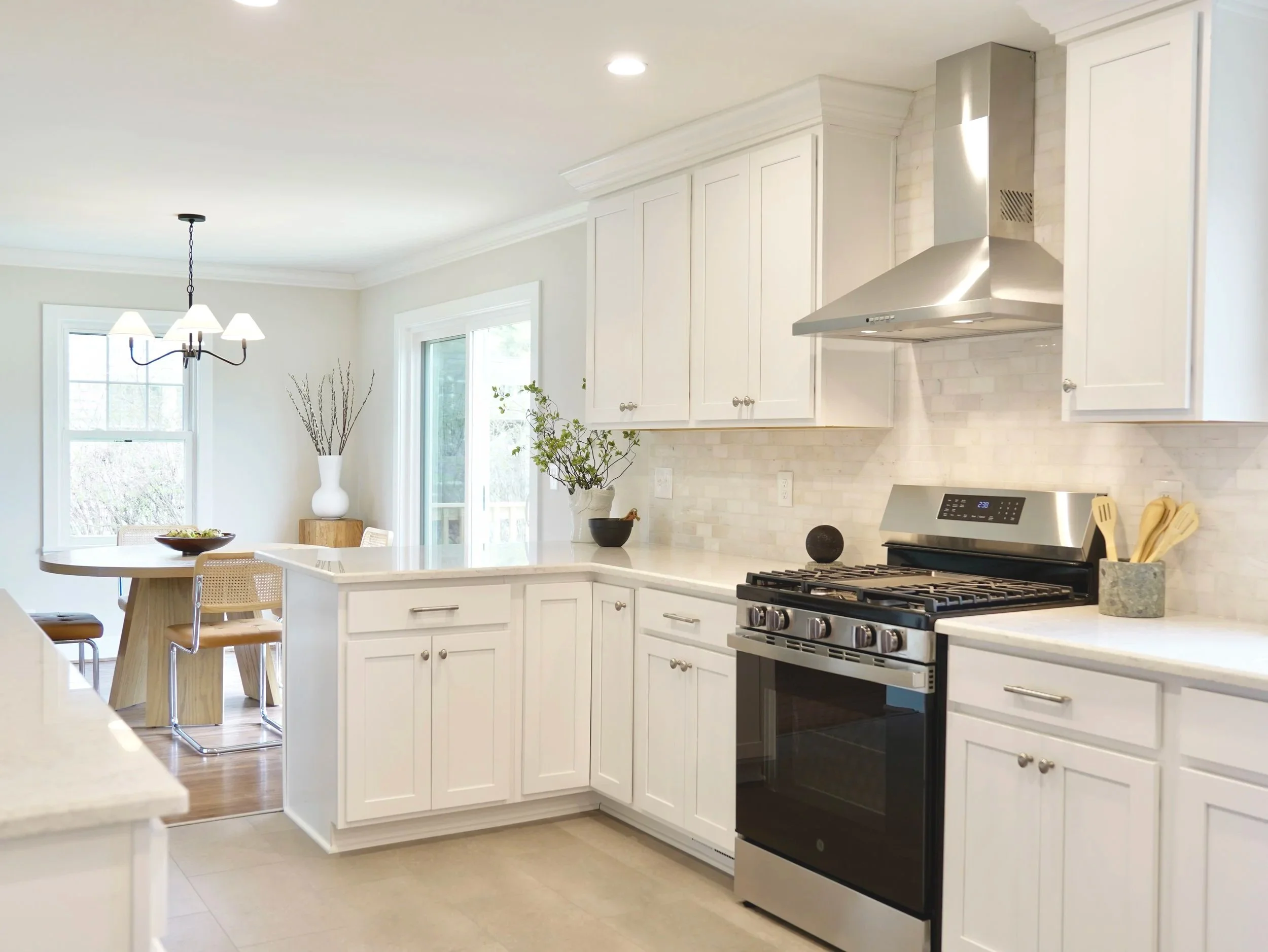 Kitchen with white cabinets, stainless steel stove and range hood, granite countertops, dining table with chairs, large windows, greenery outside, decorative plants and vase inside.