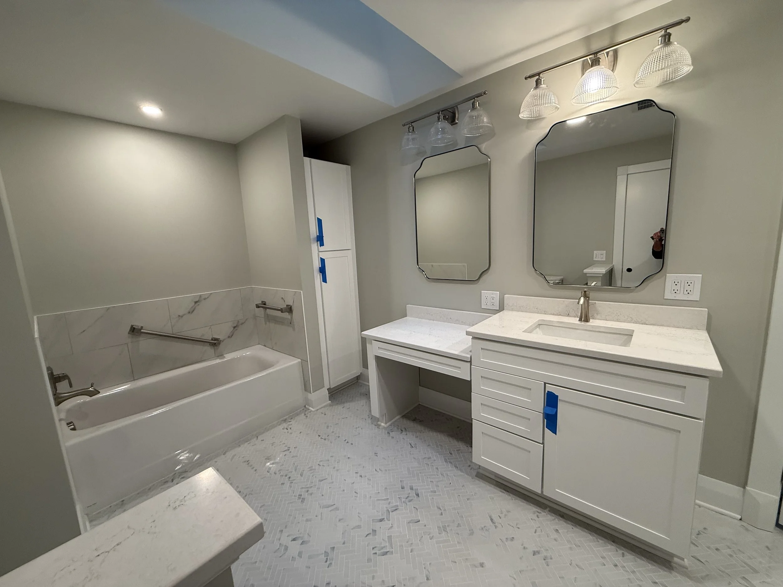 A modern bathroom with a bathtub, a double vanity with two mirrors, and a shower area with two grab bars. The room has light-colored walls, fixtures, and a patterned tile floor. There is a light fixture above the mirrors and various electrical outlet