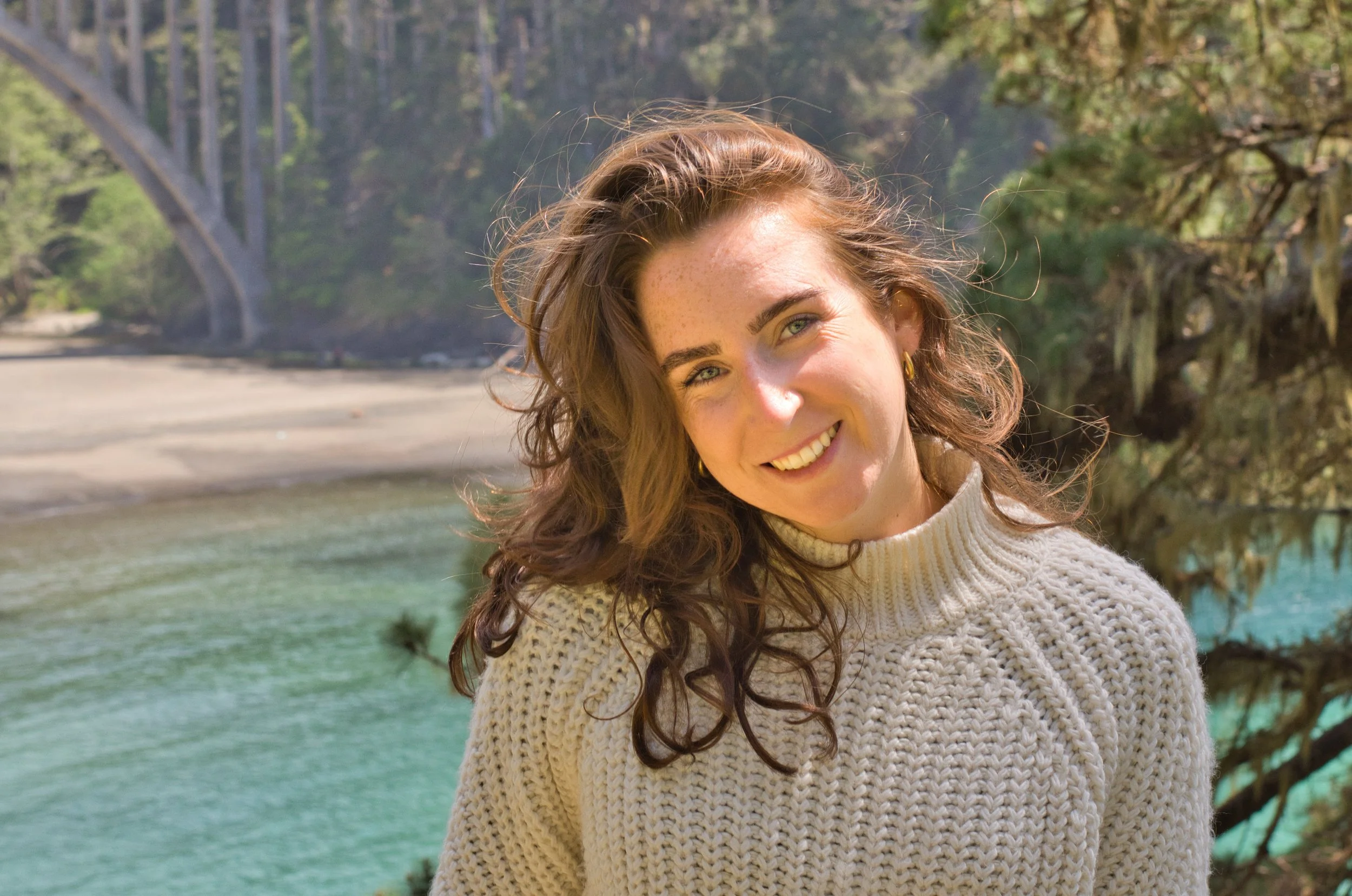 A young woman with long curly brown hair smiling outdoors near a body of water, wearing a cream-colored knit sweater, with trees and greenery in the background.