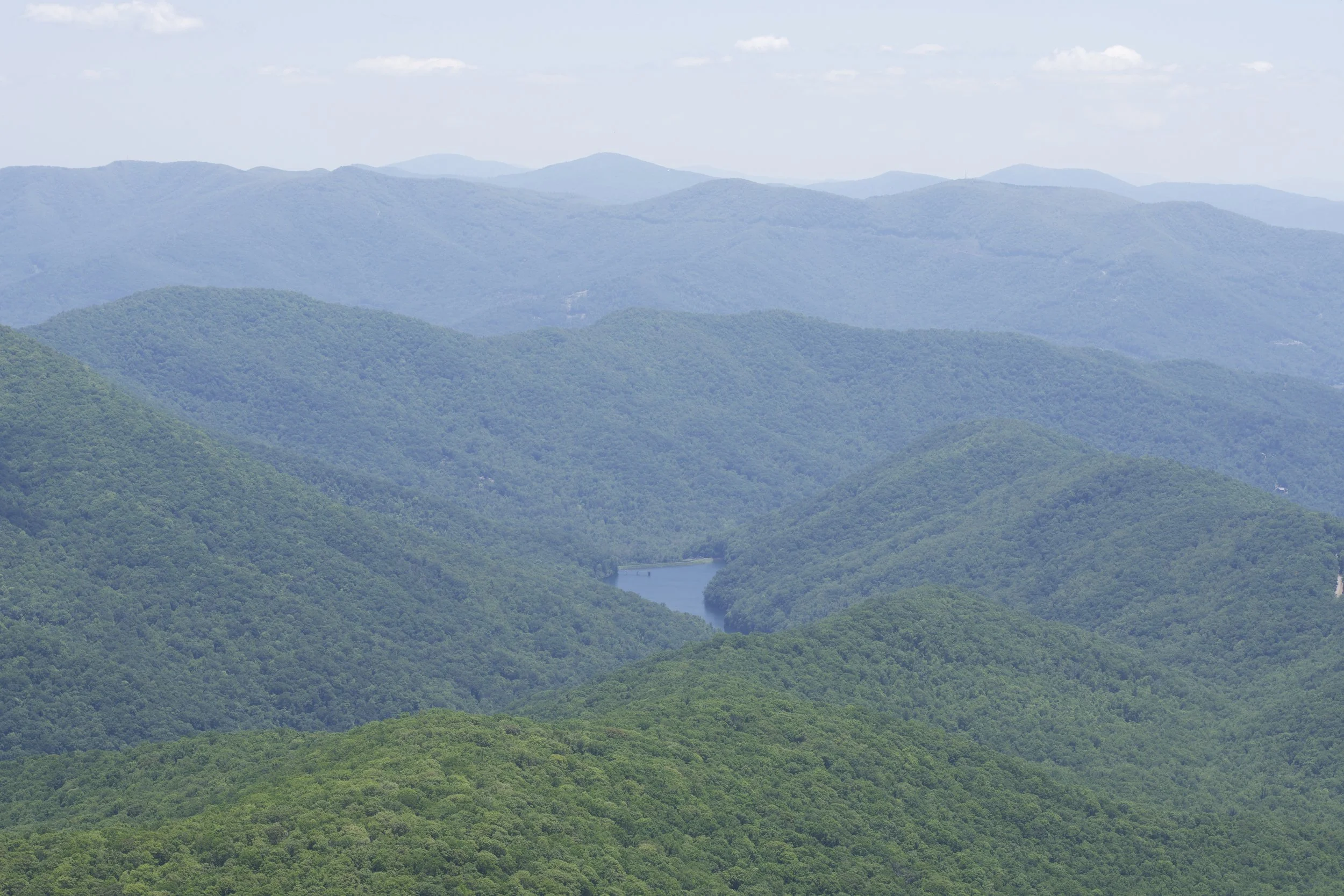A landscape of rolling green forested hills with a river running through the valley, under a partly cloudy sky.
