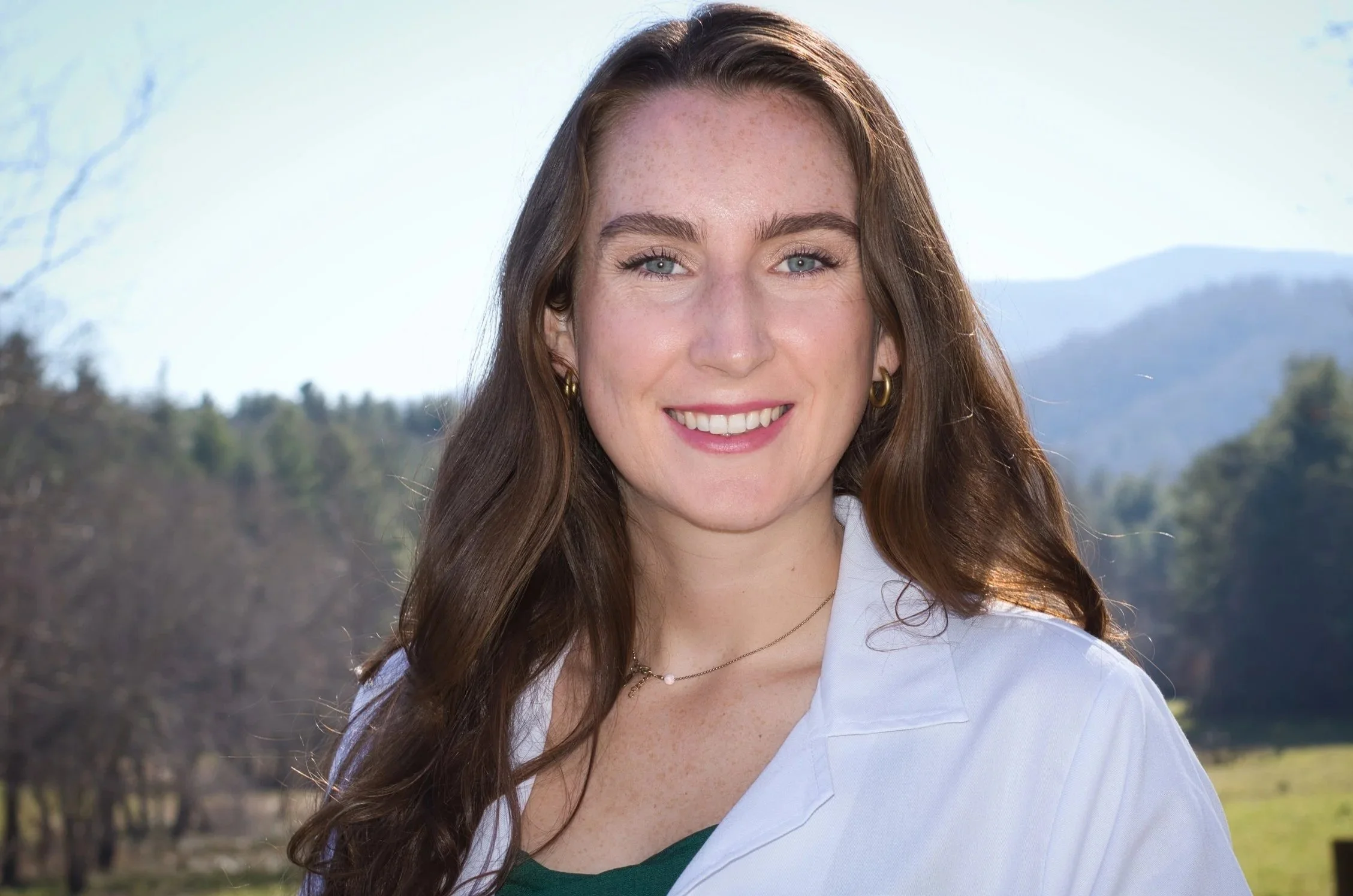 Close-up of a young woman with long brown hair, blue eyes, and earrings, smiling outdoors against a background of trees and mountains.