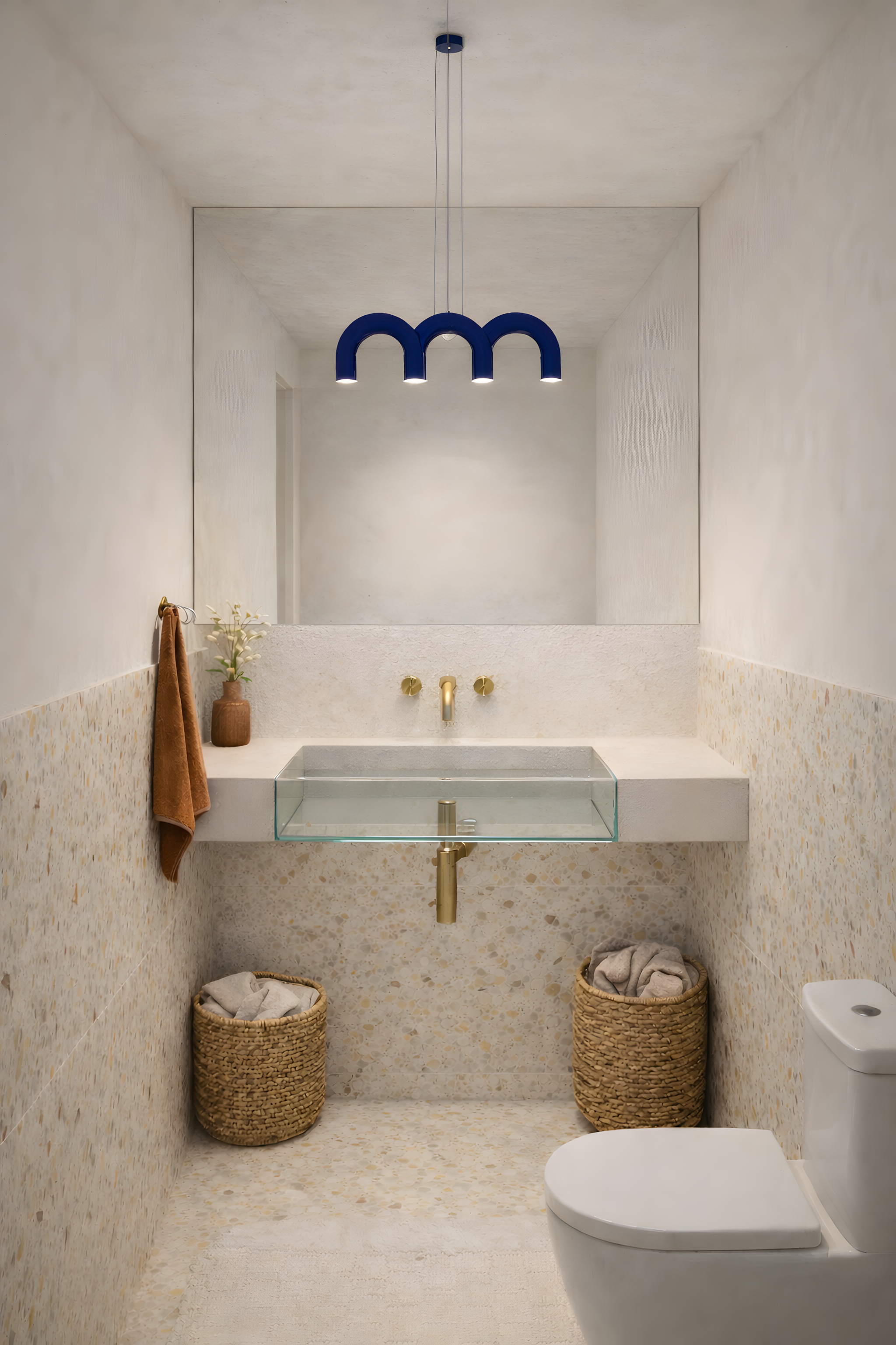 Compact guest bathroom with terrazzo surfaces, brass fixtures, glass sink and blue sculptural pendant lighting in Milan apartment interior by Zajal.