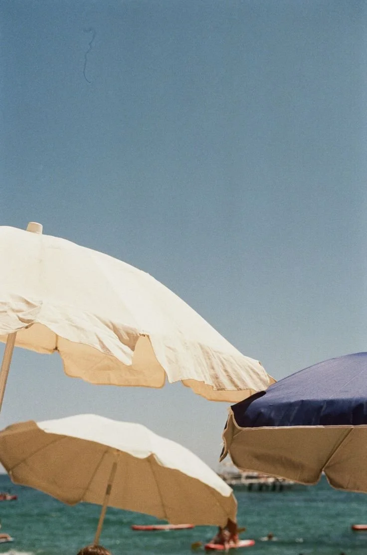 Mediterranean beach scene with cream and blue parasols against a bright coastal sky and deep blue sea.