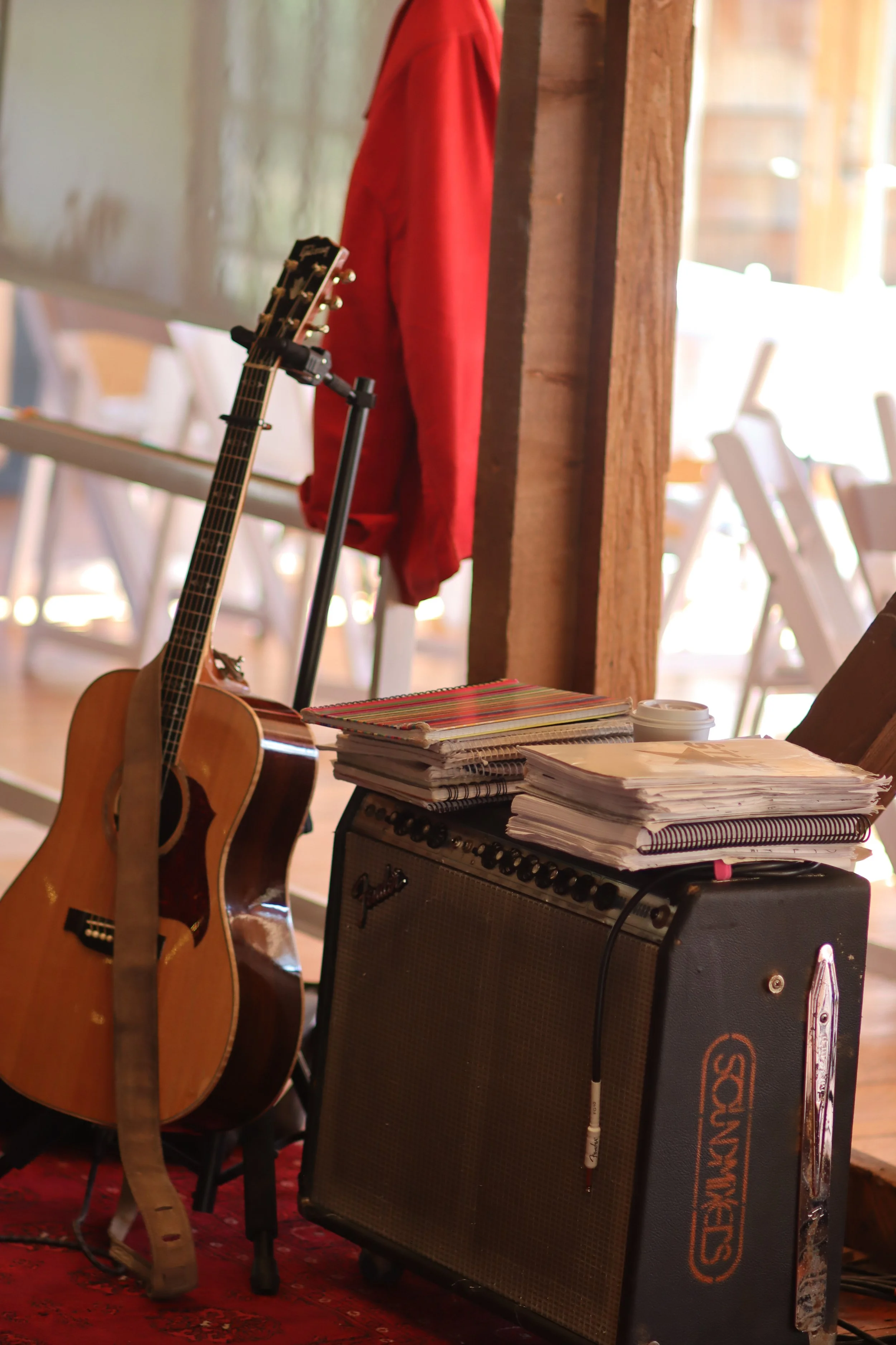An acoustic guitar leaning against a black amplifier, with stacks of notebooks and a disposable coffee cup on top. In the background, a red jacket hangs on a wooden post, and chairs are visible in a bright, indoor space.