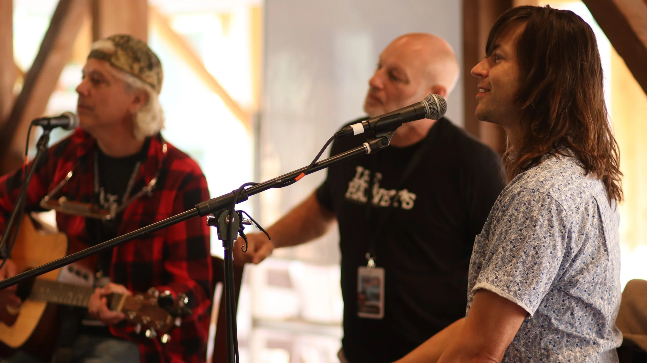 A group of three people sitting with microphones, one of whom is playing a guitar and wearing a red plaid shirt, another in a black shirt, and a woman in a blue floral blouse, all smiling or serious, indoors with natural light.