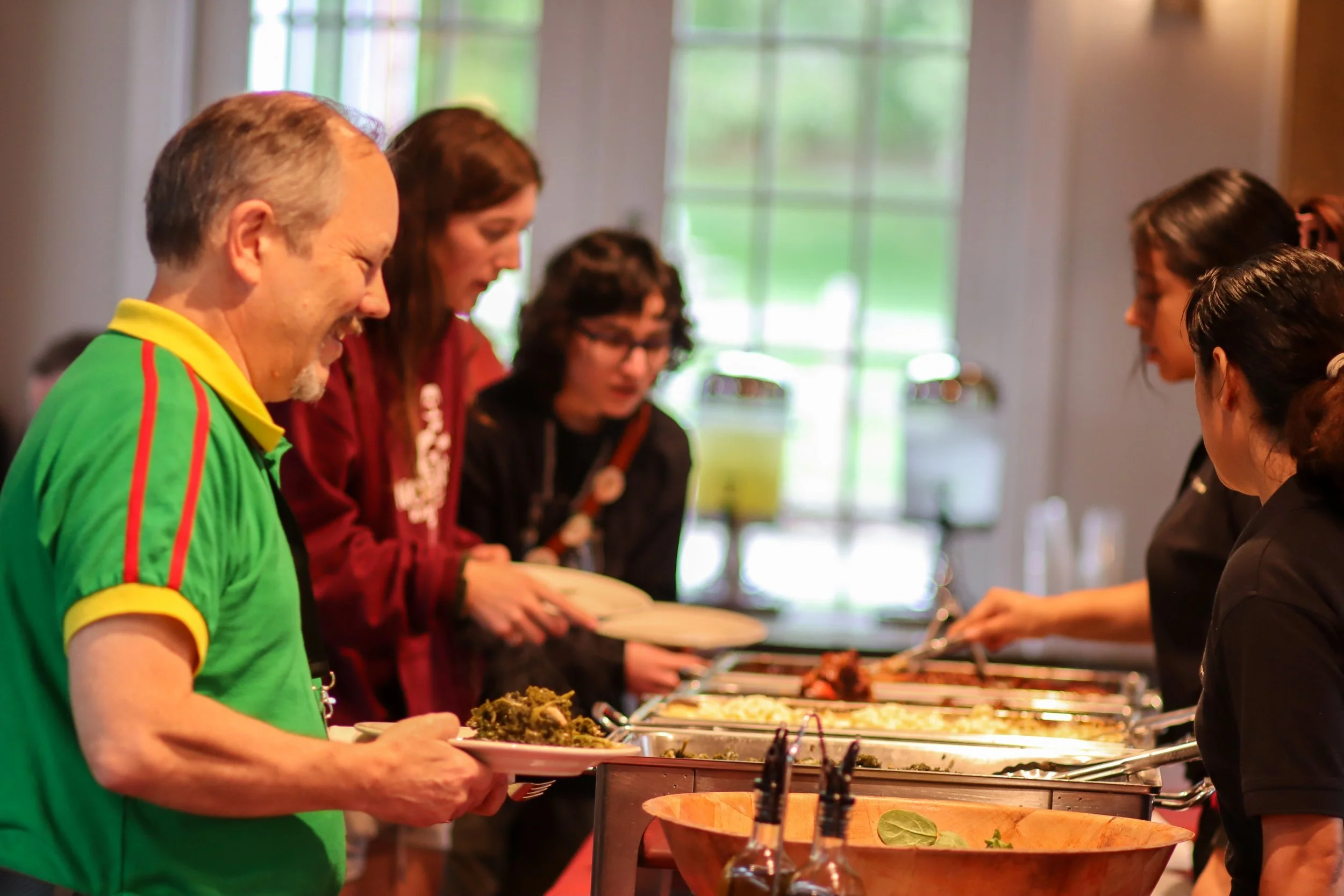 People serving themselves food from a buffet with several dishes in chafing dishes in a well-lit room with large windows.