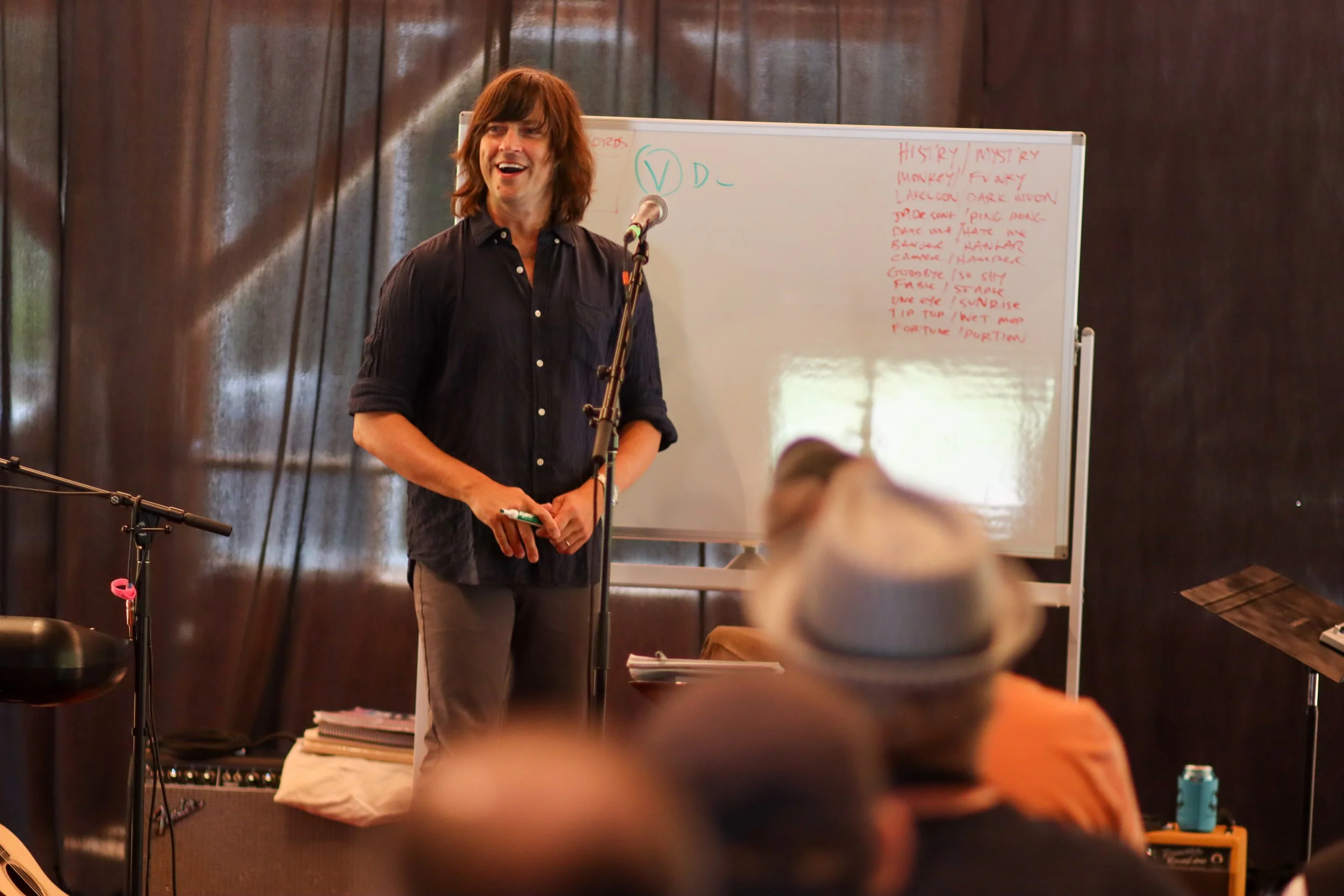 A man with long brown hair, wearing a black shirt, smiling and standing in front of a whiteboard with handwritten notes, in a room with dark wood-paneled walls, speaking into a microphone.