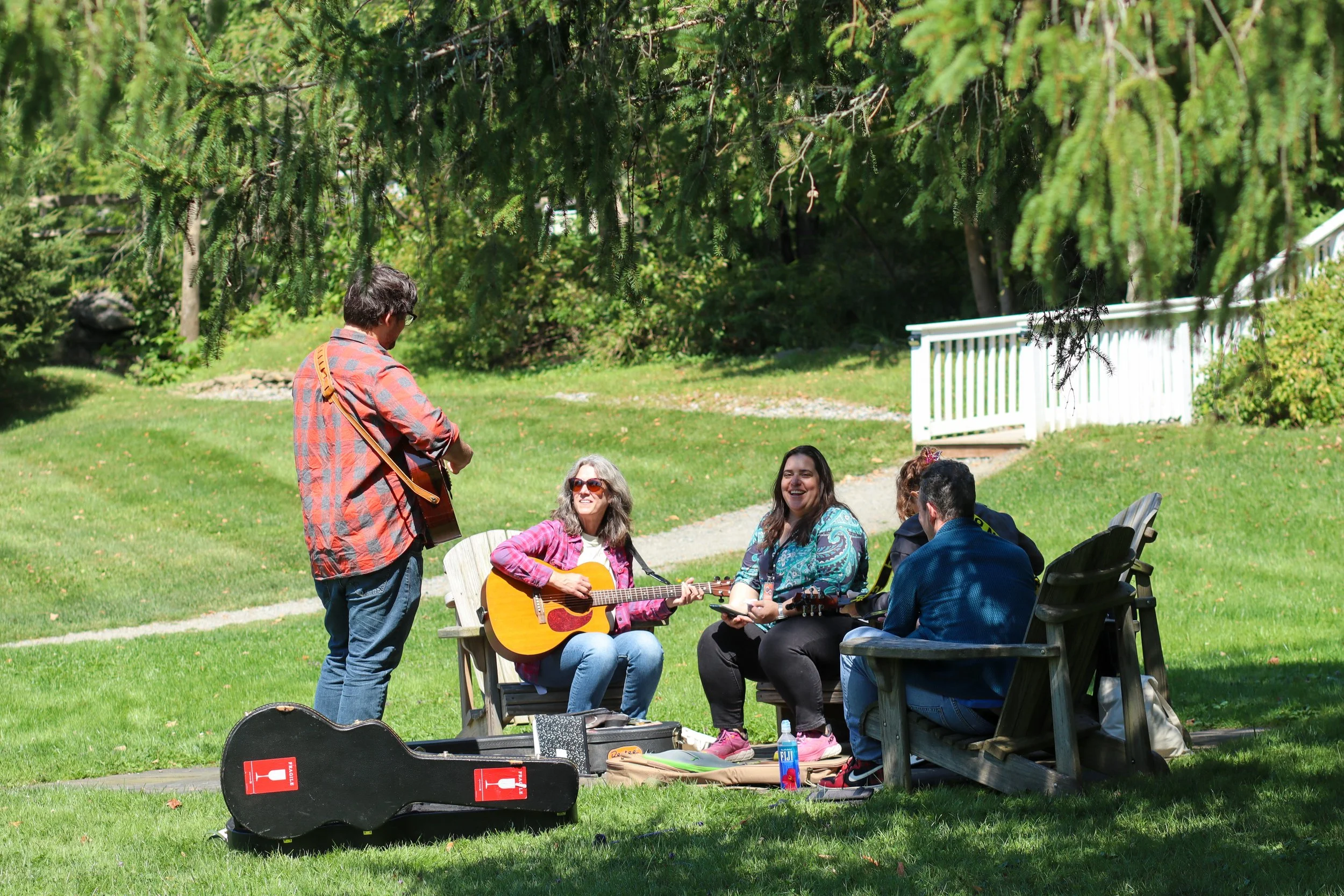 Group of five people sitting on benches outdoors in a grassy area, with one person playing guitar and another appearing to sing or listen, under green trees with sunlight filtering through.