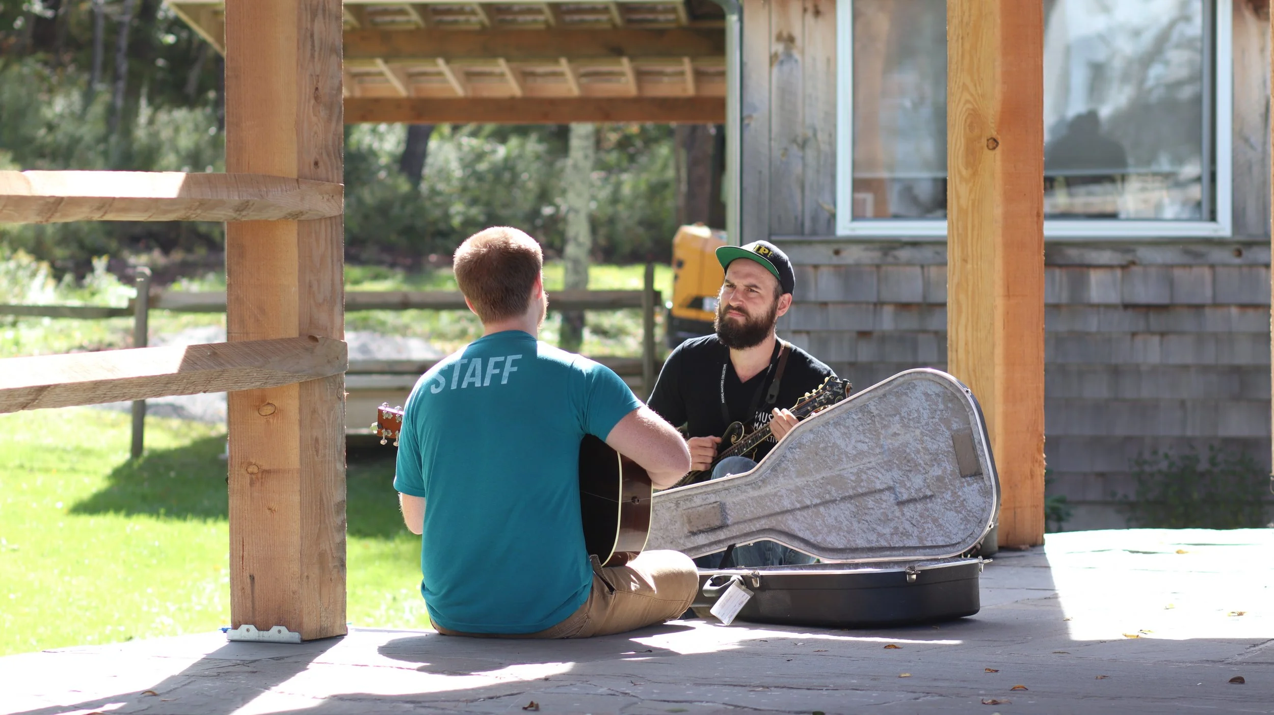 Two men sitting outdoors under a wooden structure, one playing guitar and the other listening.