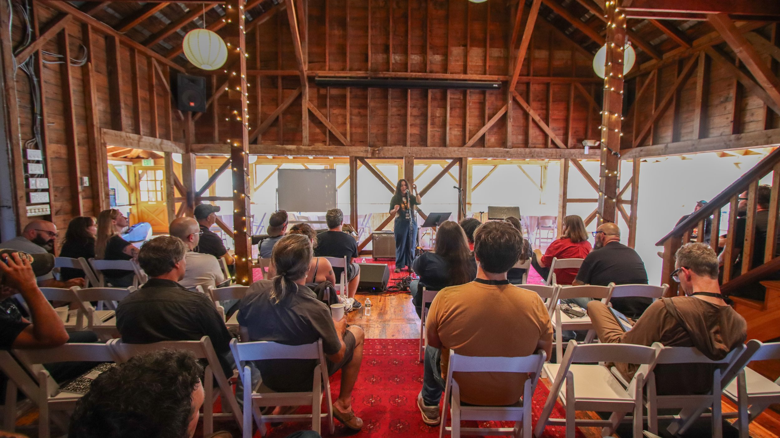 A woman on stage speaking into a microphone at a concert or event in a rustic wooden venue with string lights. Audience members seated facing the stage listen attentively.