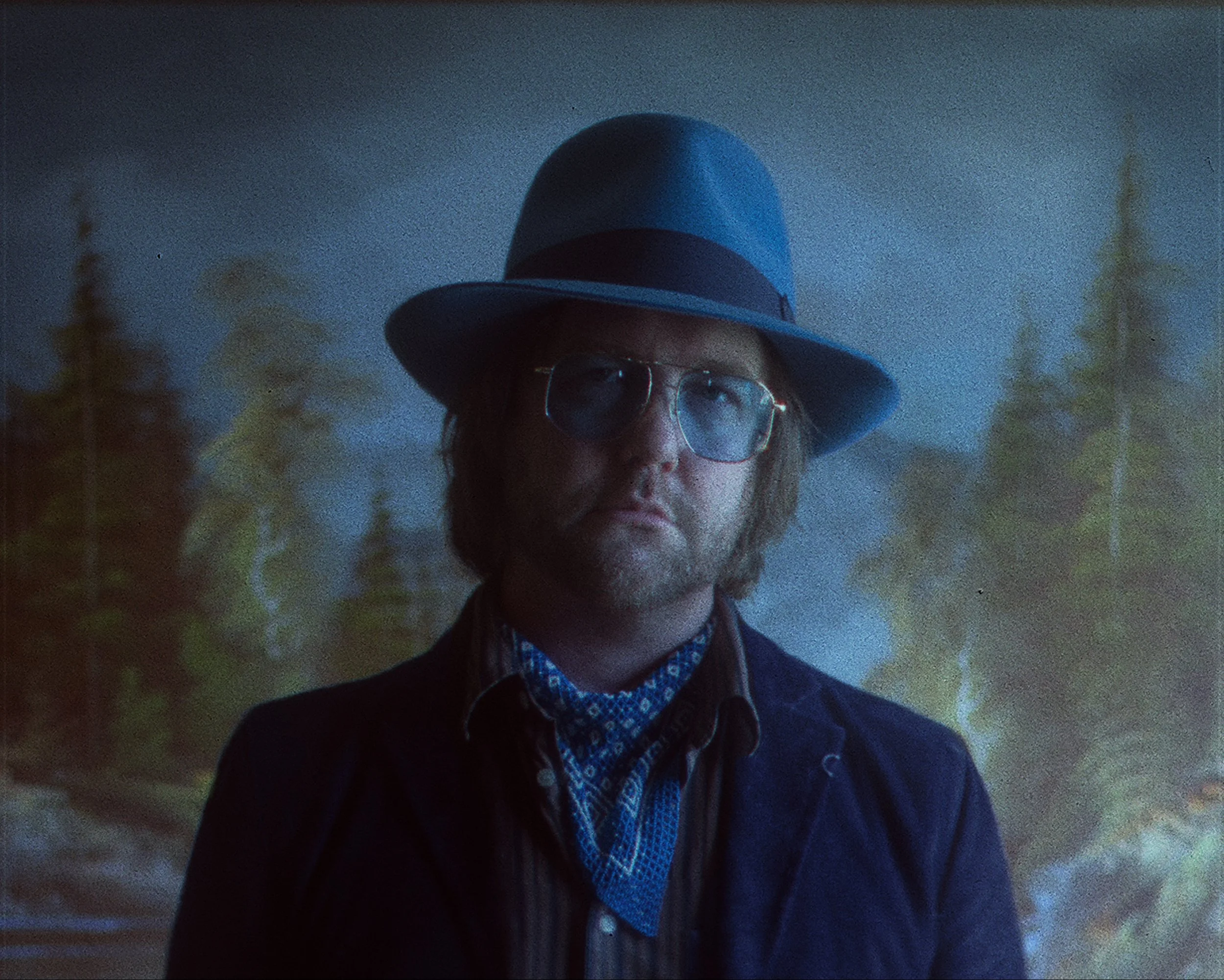 A person with glasses and long hair wearing a blue wide-brimmed hat and a patterned shirt, standing outdoors during dusk or dawn.