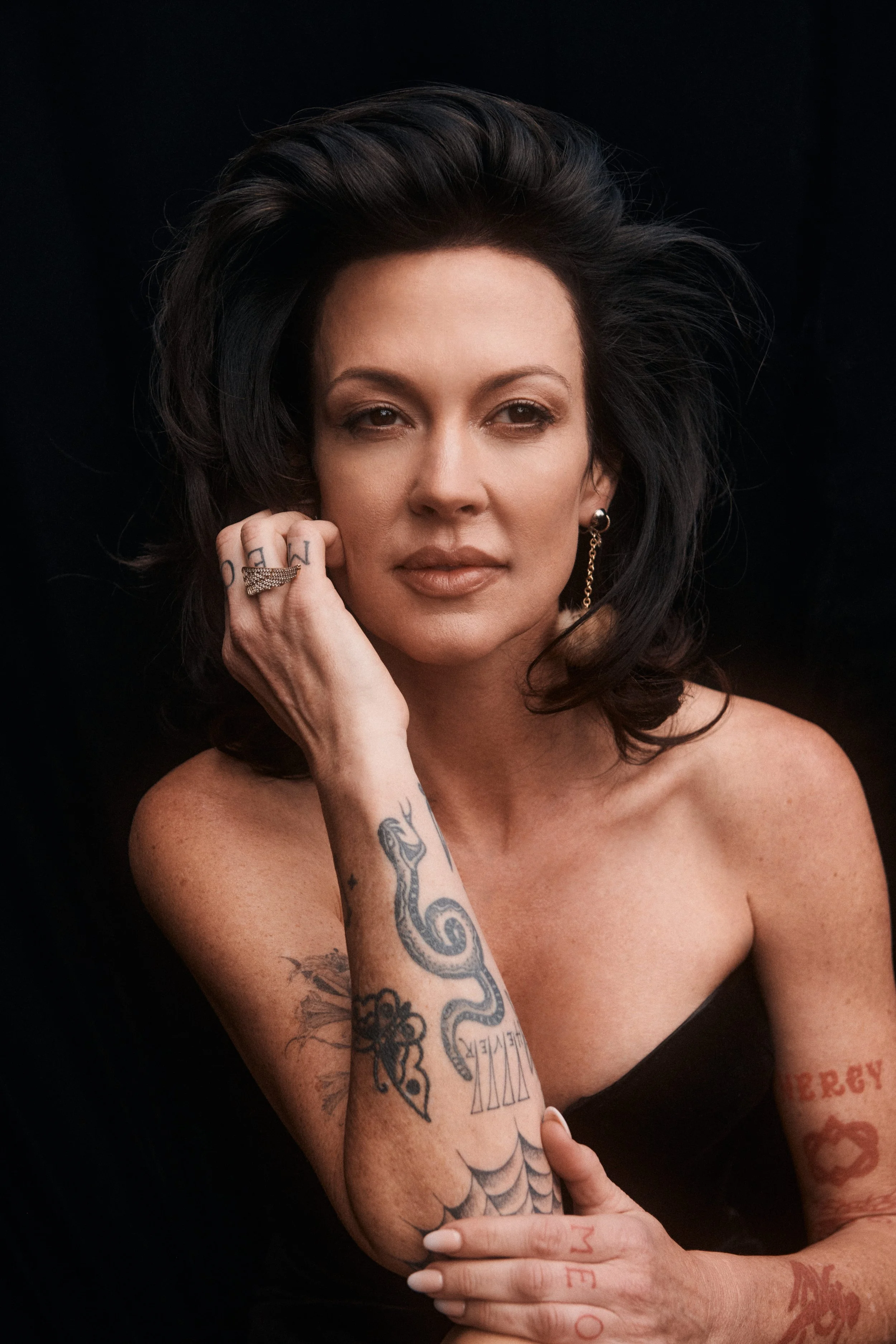Portrait of a woman with dark, voluminous hair, tattoos on her arms and chest, wearing earrings and a ring, against a black background.