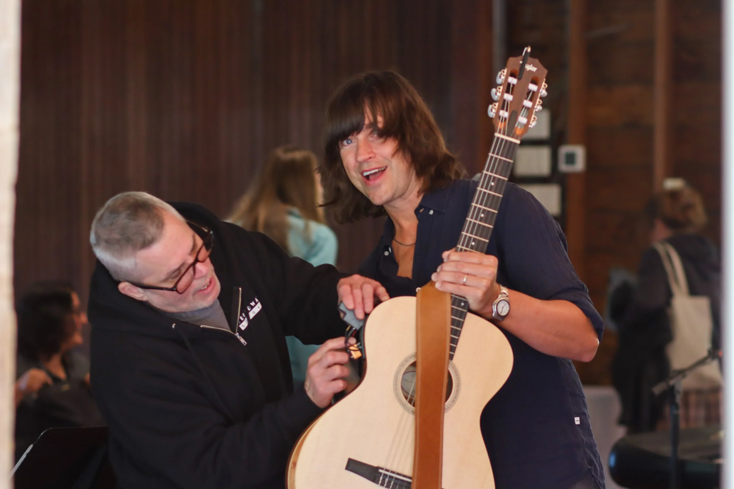 A man with gray hair and glasses, and a woman with long dark hair, are joyfully playing a guitar together in a social setting. The woman is holding the guitar and smiling at the man, who is also smiling and appears to be enjoying the moment.