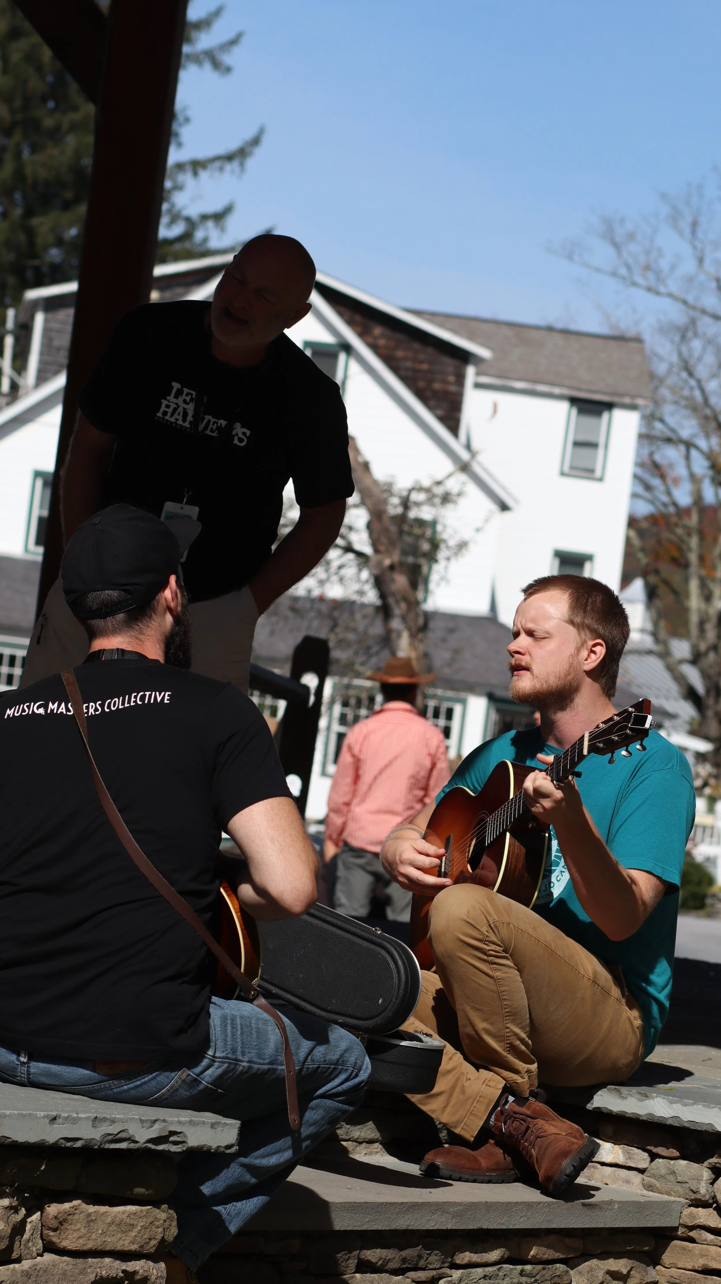 Three men with guitars performing outdoors; one sitting on a stone ledge, another sitting on the ground, and the third standing.