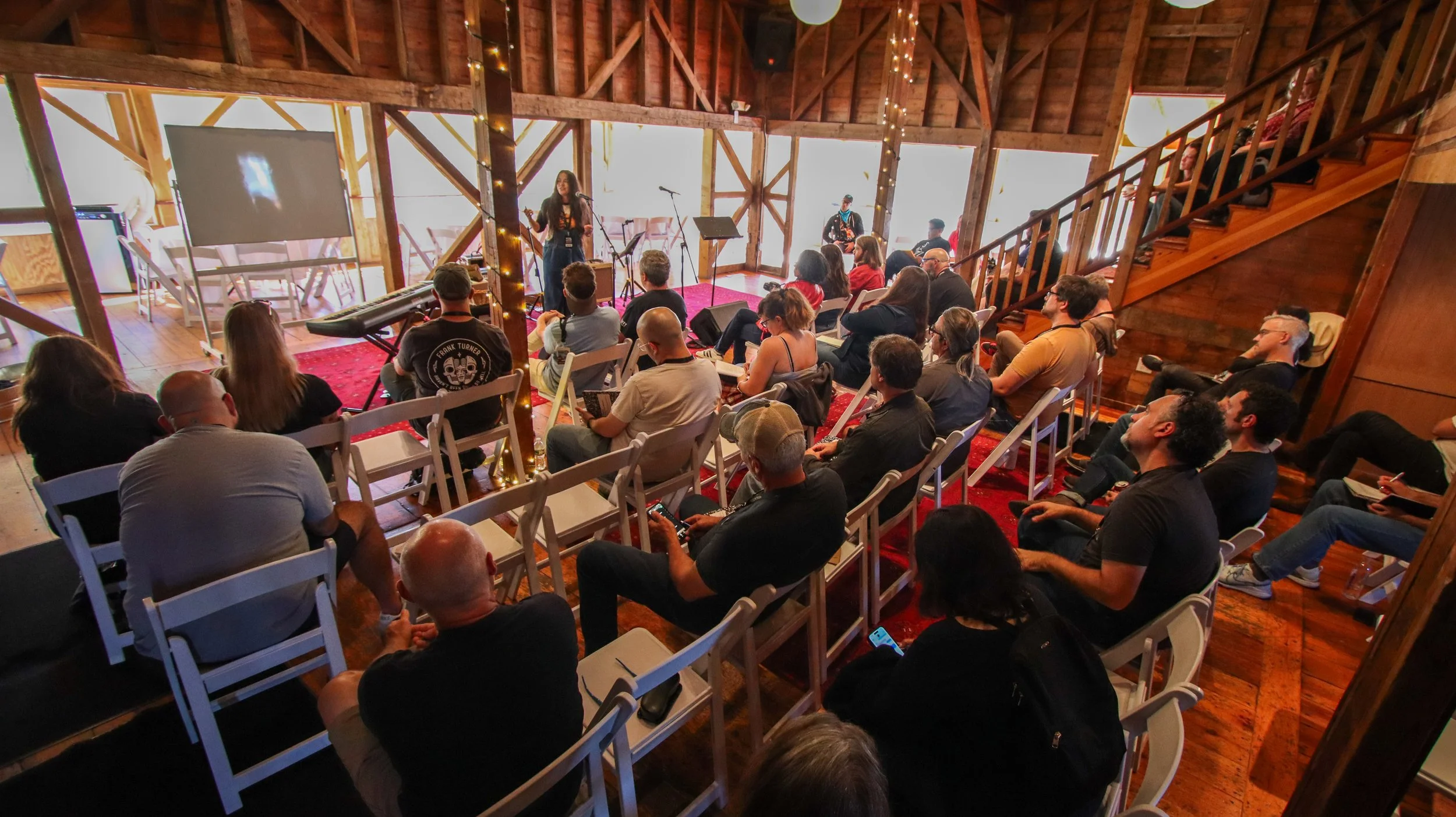 People gathered in a rustic wooden venue watching a live musical performance on a small stage, with some seated and some standing around.