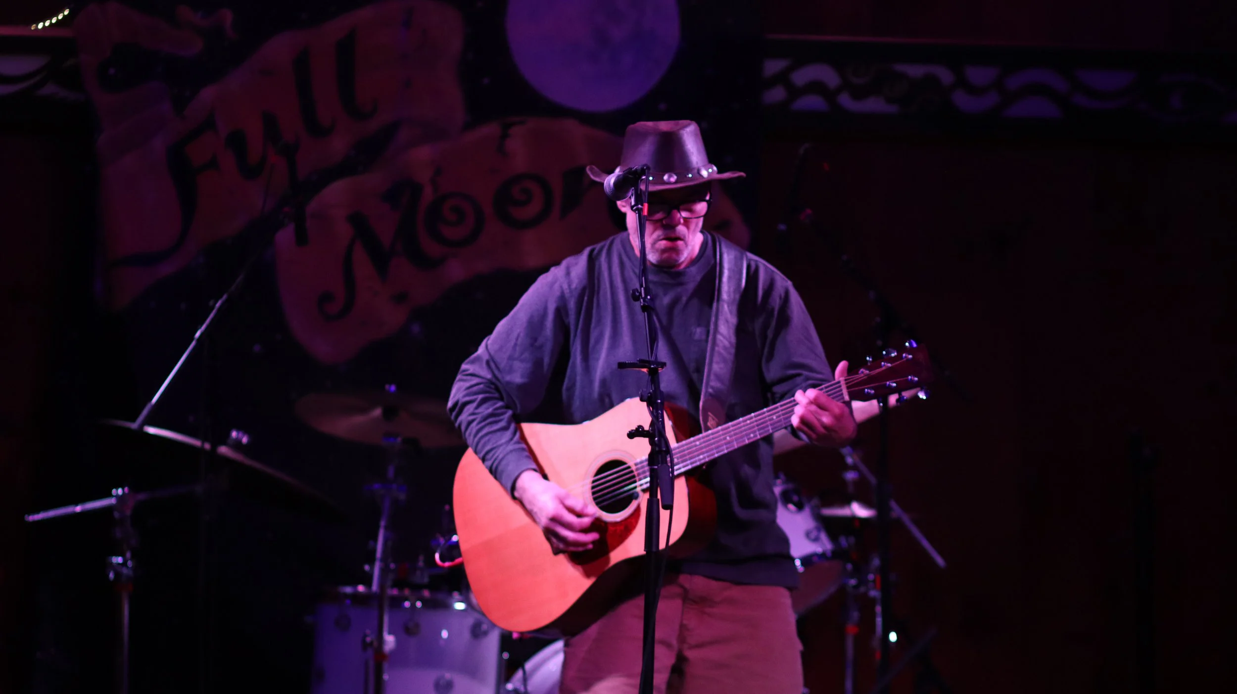 A man wearing a cowboy hat and glasses playing an acoustic guitar on stage with purple lighting, a drum set in the background, and a colorful mural or banner with the word "Funk" behind him.