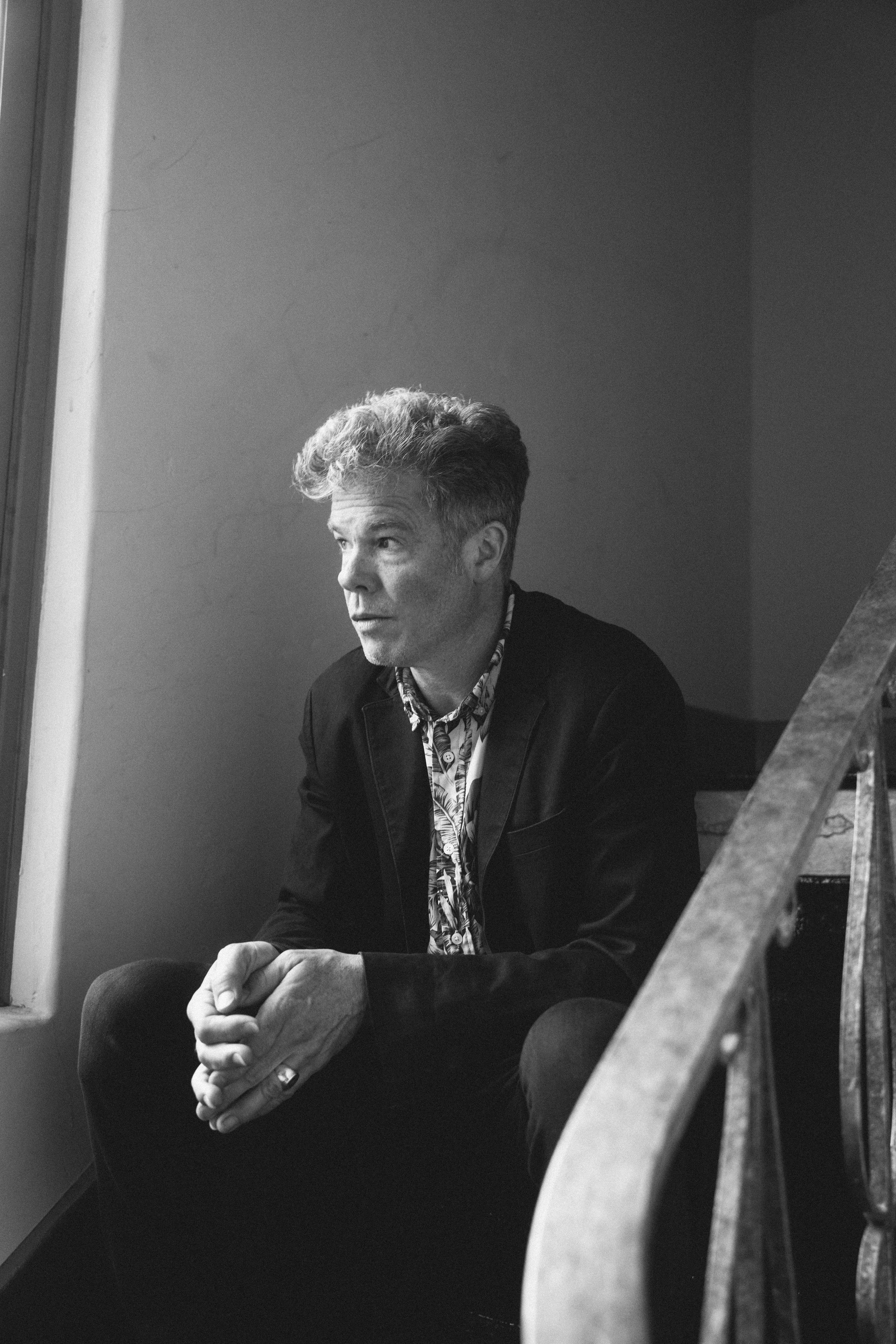 Black and white photo of a man sitting on wooden stairs near a window. He has curly hair, wears a blazer, and has his hands clasped. The photo captures a contemplative expression.