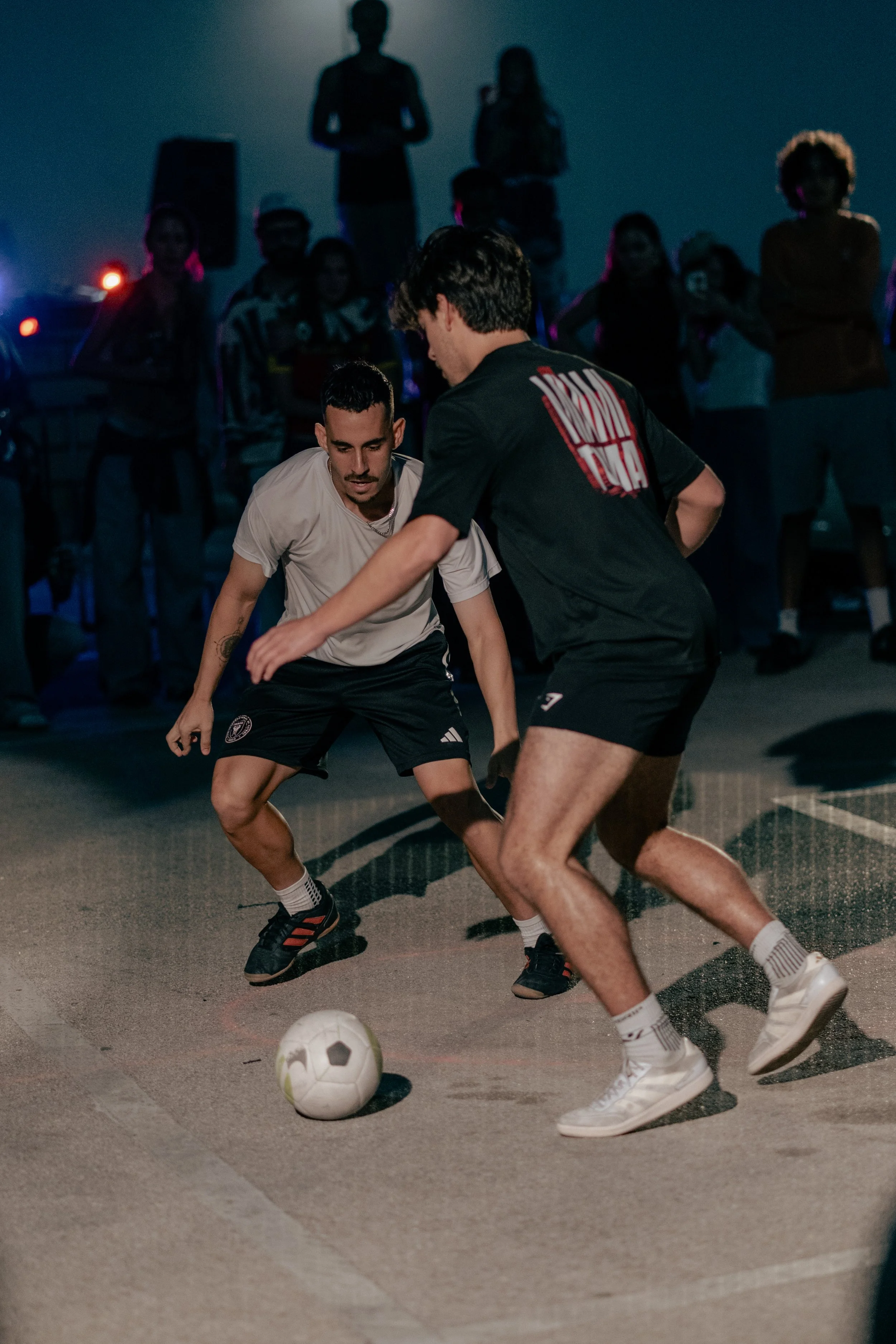 Two young men playing soccer on a court at night with a group of spectators watching.