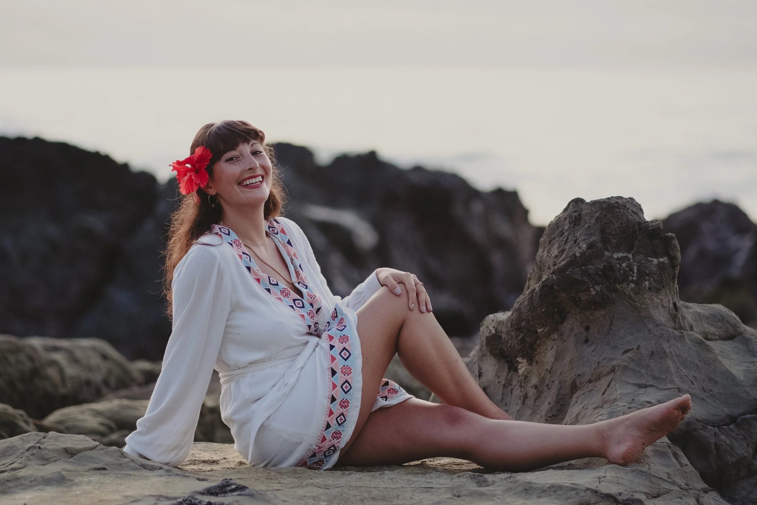 Woman in white embroidered dress with a red flower in her hair sitting on rocks at the beach, smiling at the camera.