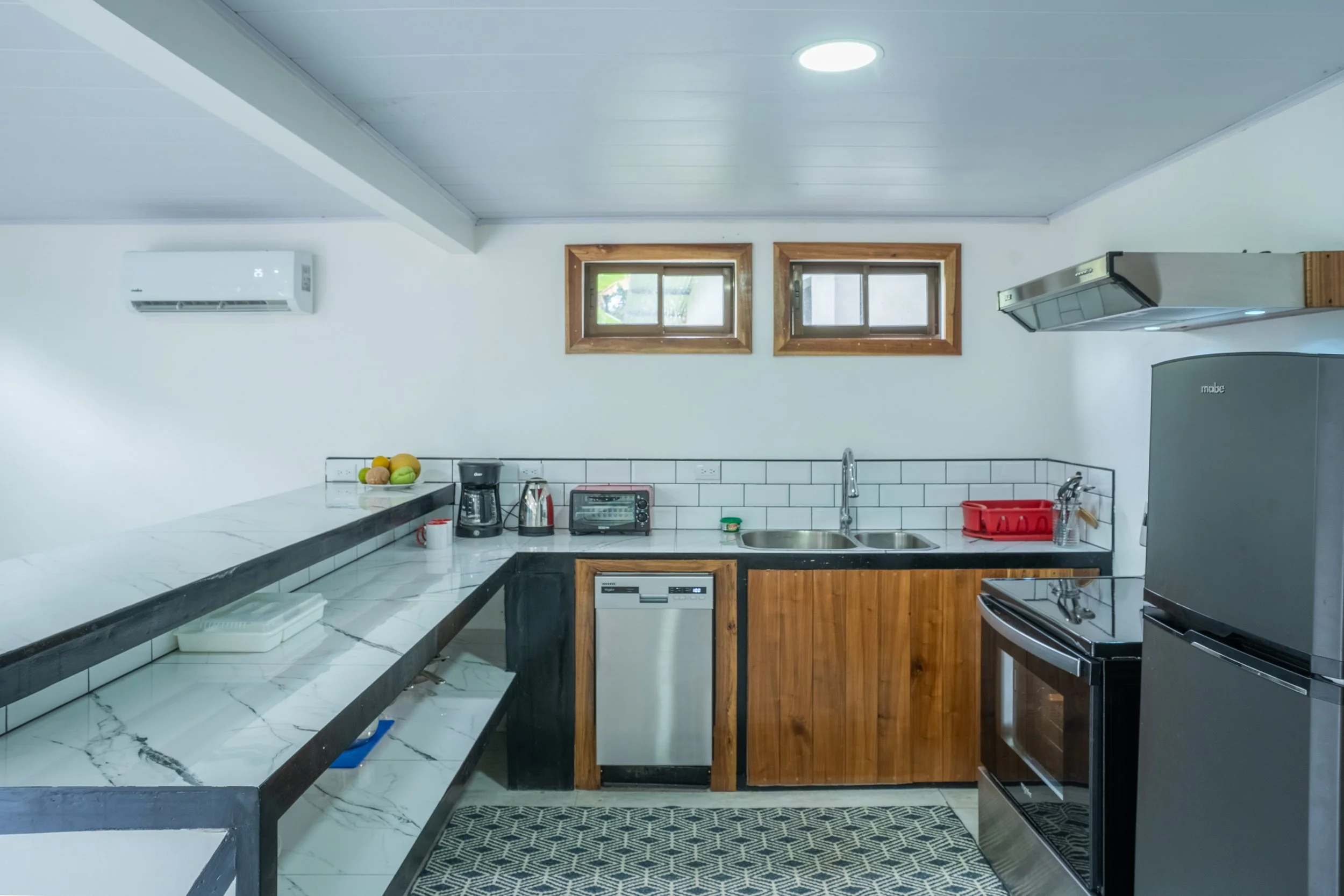 Kitchen with white walls, two small windows with wooden frames, gray refrigerator, oven, double sink, coffee maker, toaster, microwave, and various kitchen items on the counter.