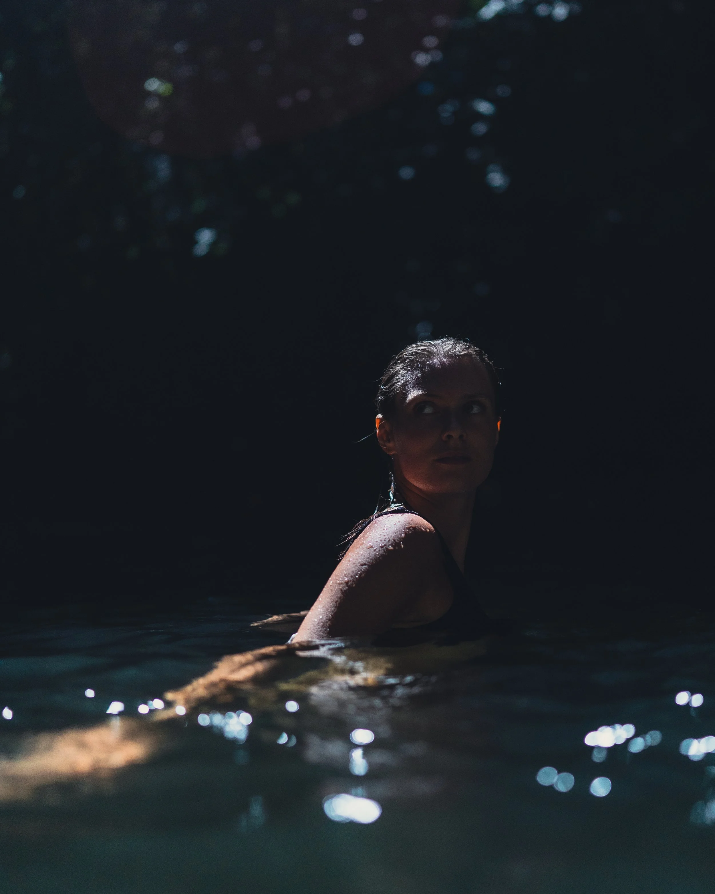 A woman with wet hair swimming in a dark body of water, with sunlight shimmering on the surface.