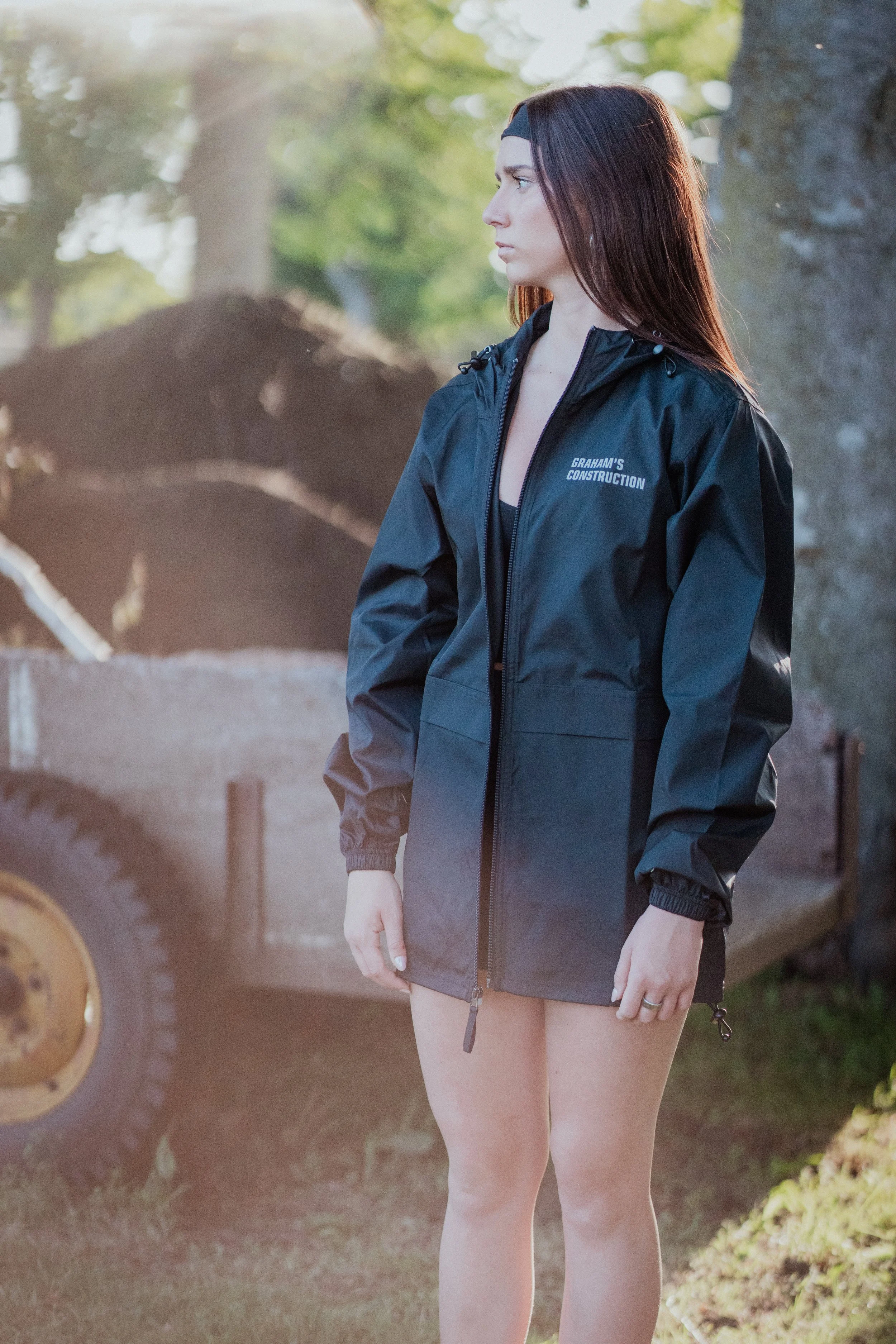 Young woman with long brown hair wearing a black rain jacket with a logo that says "Graham's Construction". She is standing outdoors near trees with a blurred background of construction equipment and dirt.
