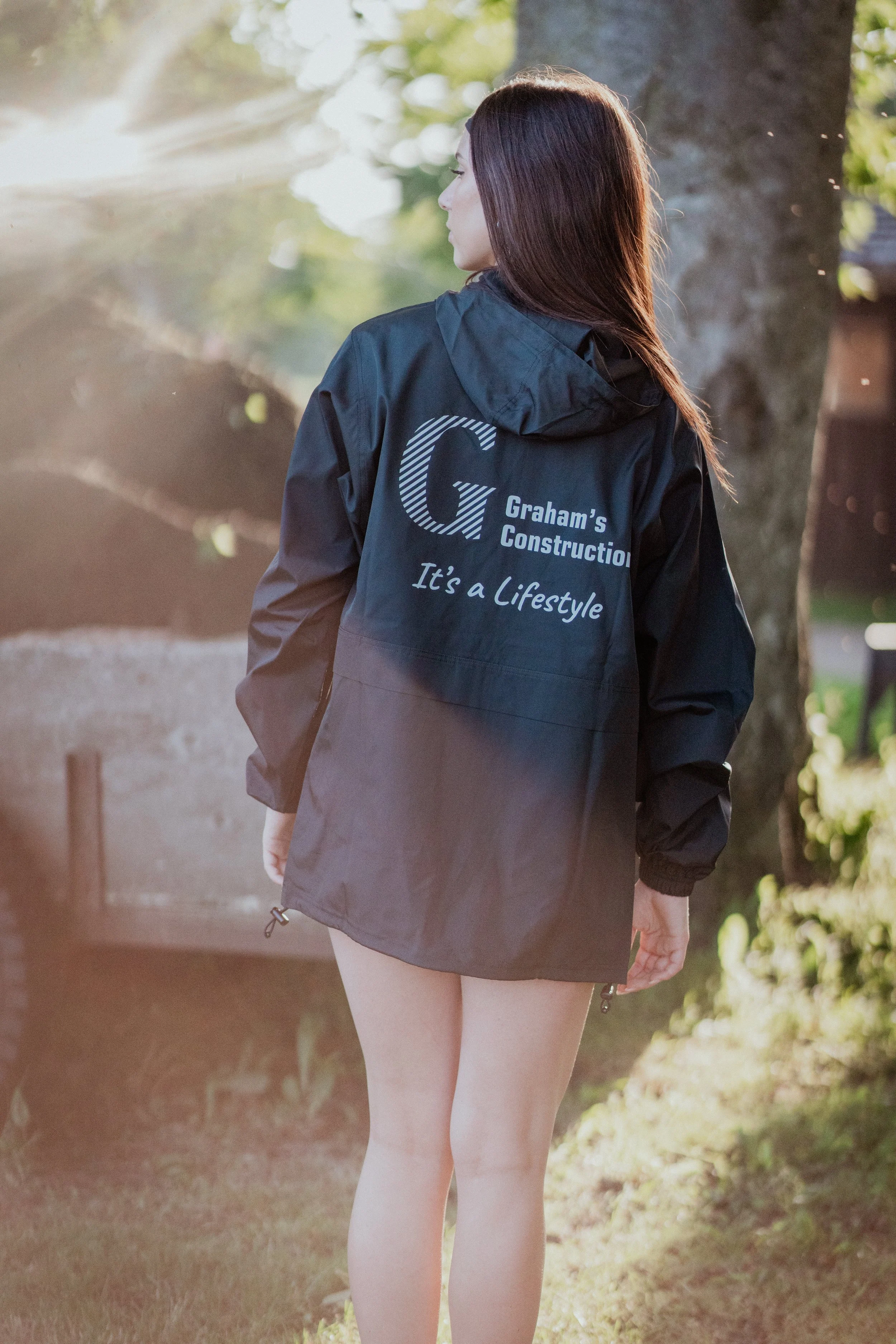 Woman standing outdoors wearing a black jacket with the logo and text for Graham's Construction on the back, surrounded by trees and sunlight.