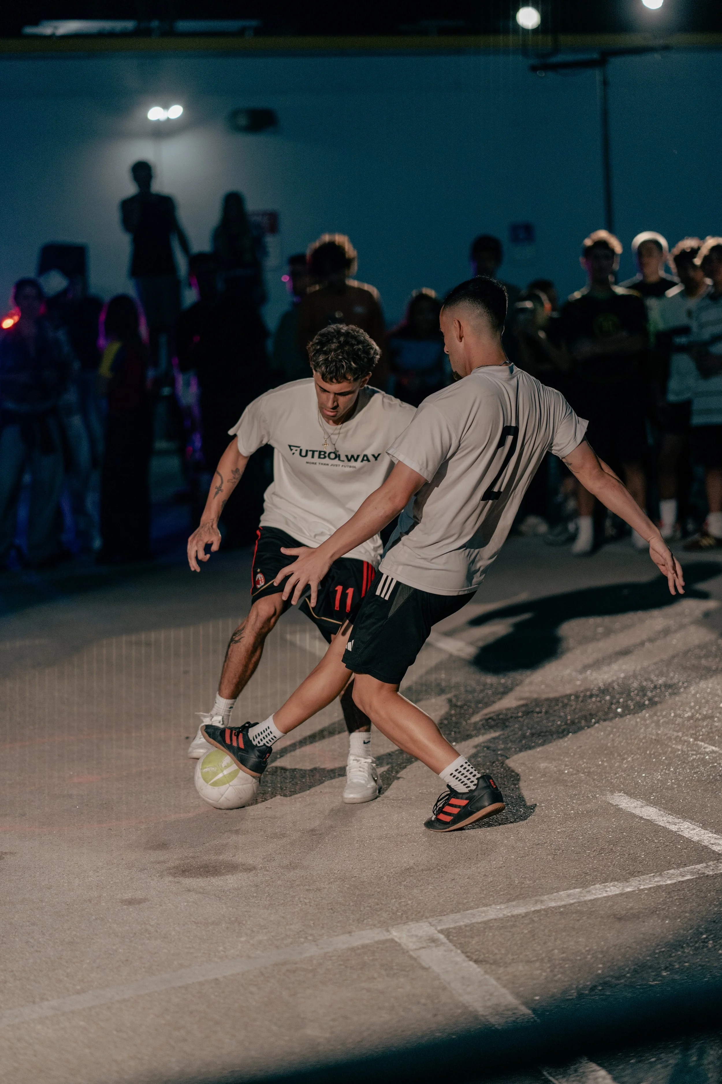 Two young men playing foot soccer on an outdoor court at night with a crowd watching in the background.
