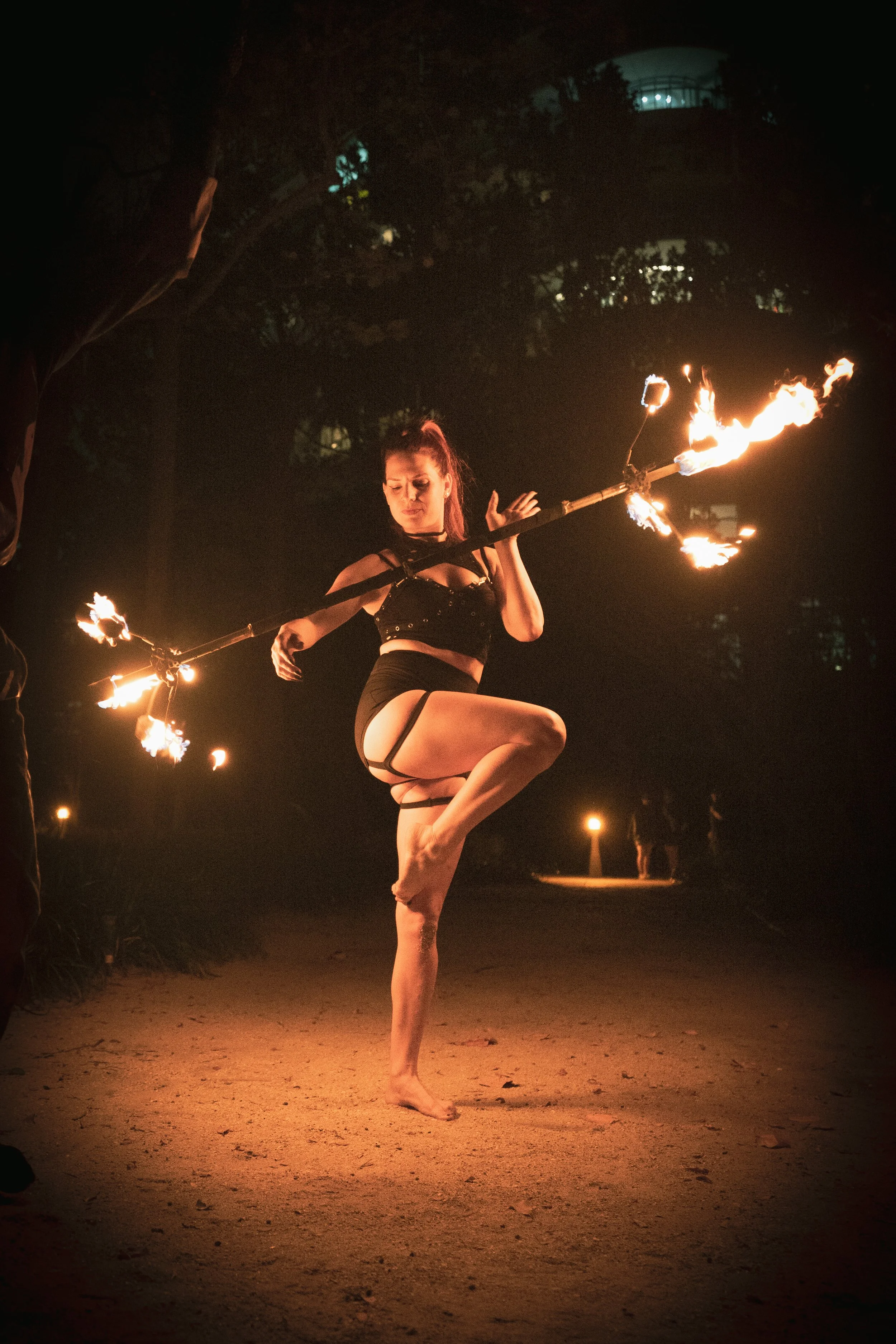 A female fire dancer performing outdoors at night, balancing on one foot while spinning a flaming staff.