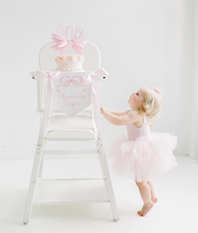 A young girl in a pink tutu dress stands on her toes, reaching for a birthday cake on a high chair. The cake is decorated with pink ruffles and topped with a pink and white bow and a 'One' cake topper. The high chair has a pink banner with the name 'Charlotte Capri' on it.