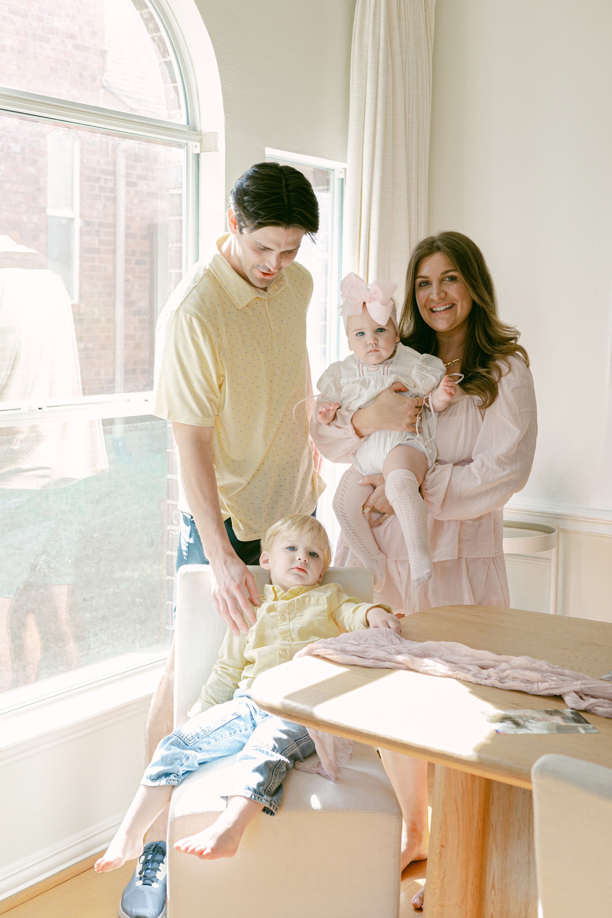 A woman with long brown hair holding a young girl with a large pink bow, standing next to a man with dark hair, and two young boys, one sitting at a table and the other standing, in a bright room with large windows and white curtains.