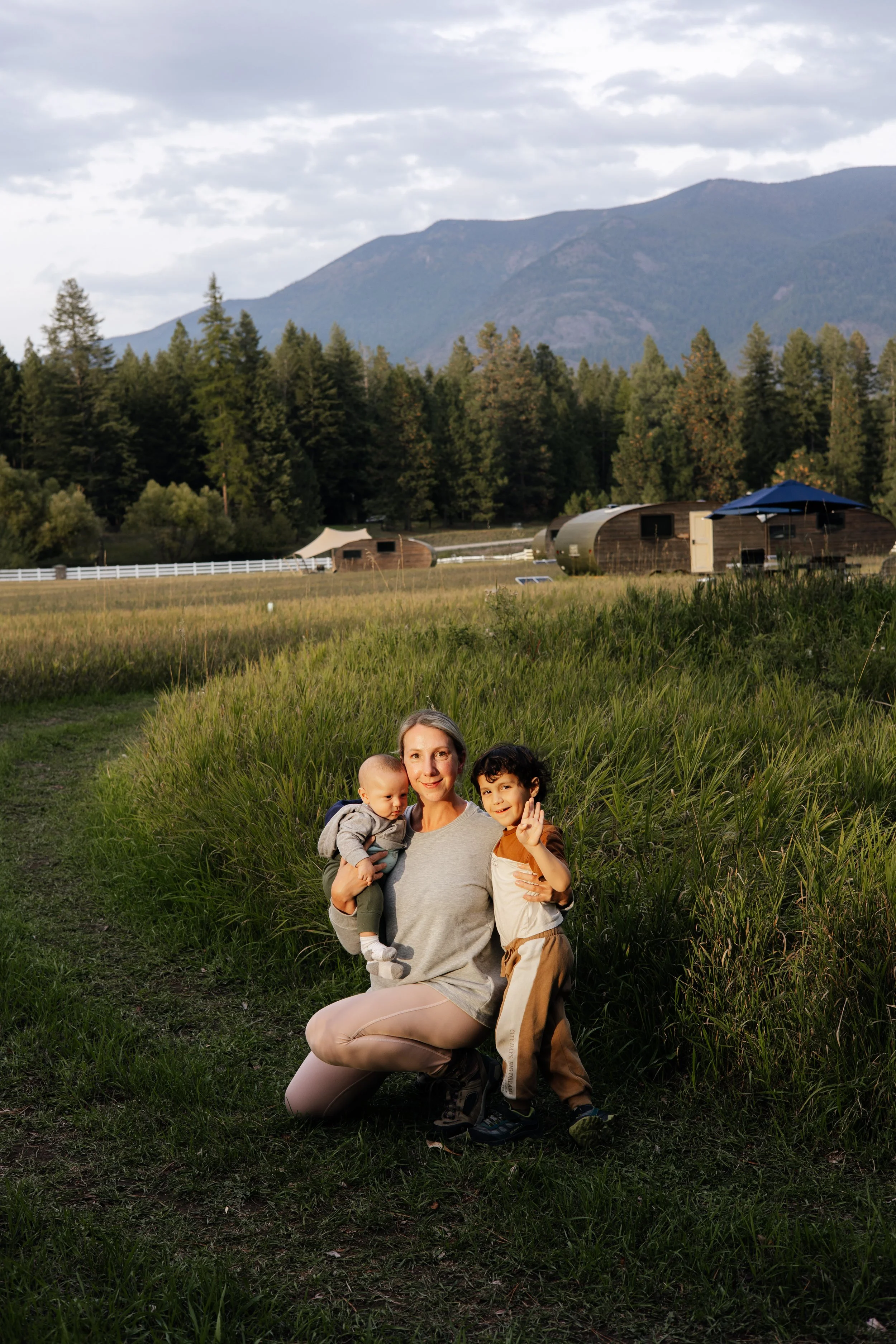 A family of three, including a woman and two children, sitting on grass with a mountain and forest in the background. The woman is kneeling, holding the younger child in her arms, and the older child is standing beside her, waving at the camera. A camping trailer and a blue umbrella are also visible.