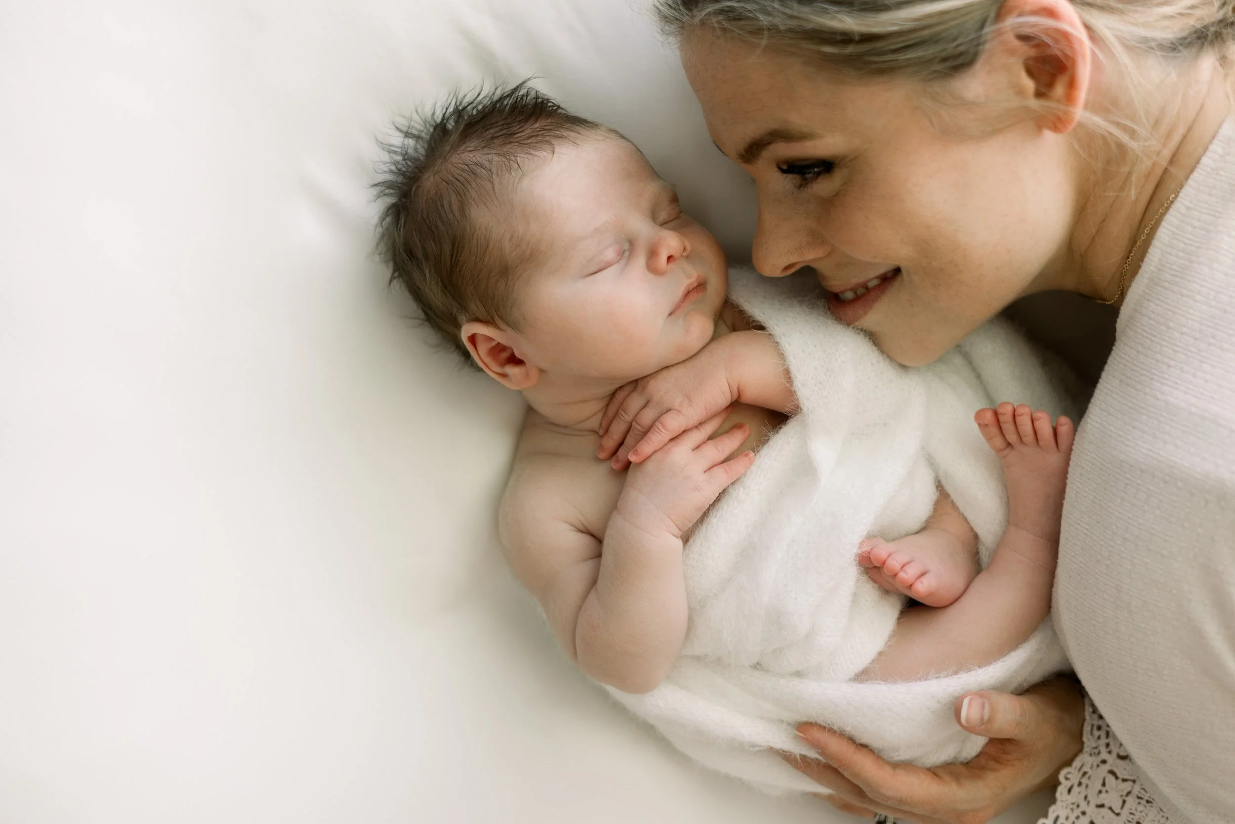 A woman holding a sleeping baby close to her face while lying down on a white surface.