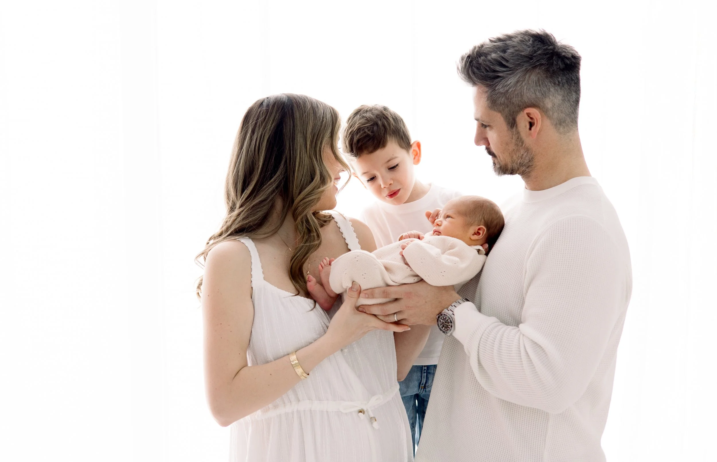 A family of four with a mother, father, a young boy, and a newborn baby, all dressed in white, standing together against a white background.