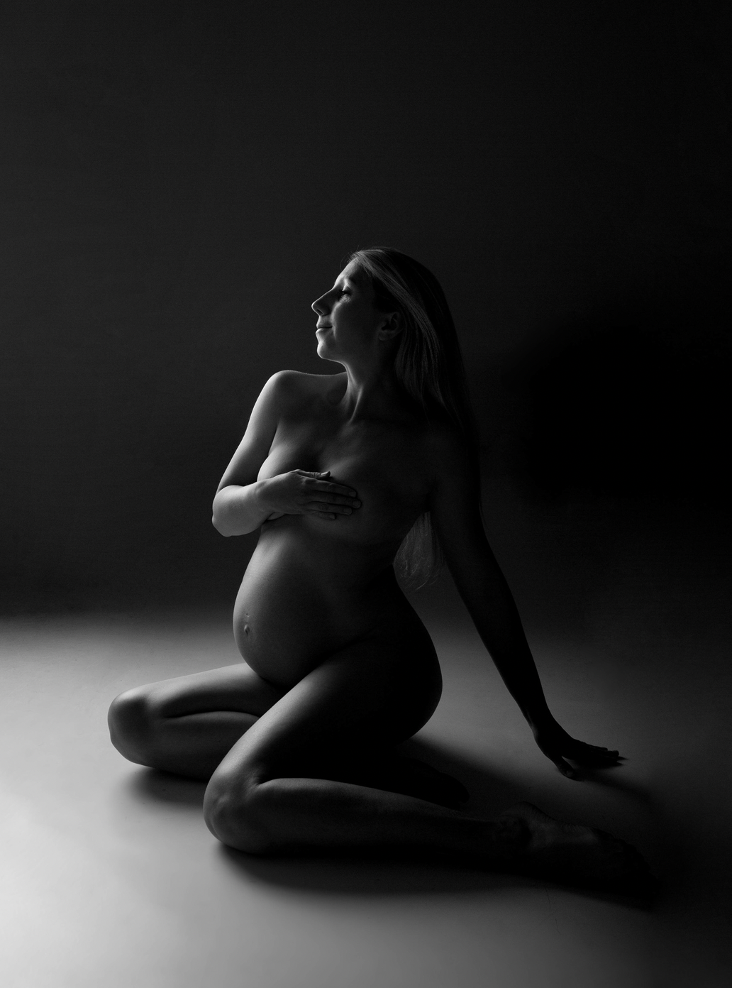 Black and white photograph of a pregnant woman kneeling on the floor, covering her breasts with one hand, with her eyes closed and head slightly tilted back. The lighting highlights her silhouette against a dark background.