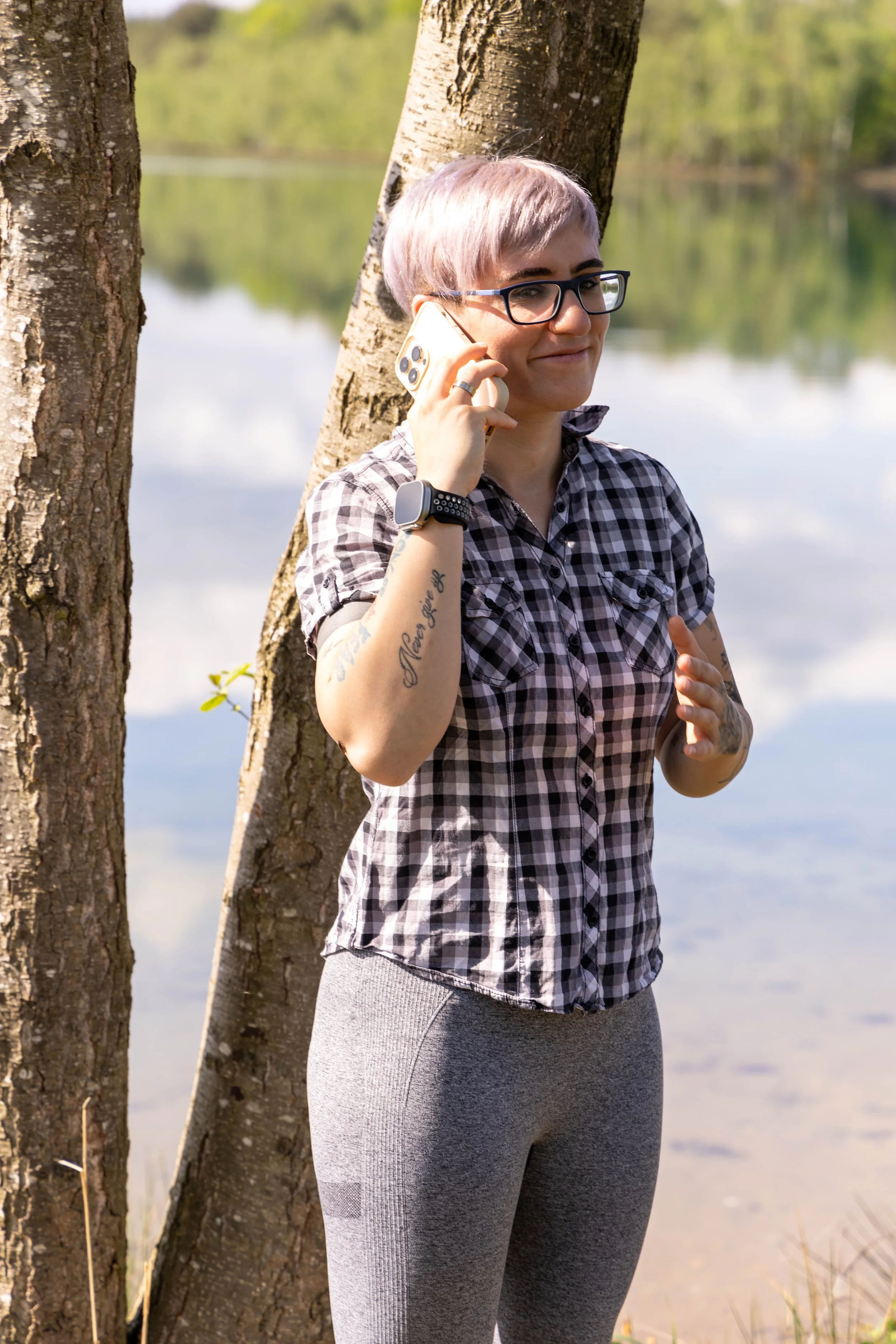 Eine Frau steht am Fluss, trägt eine Brille, ein kariertes Hemd und graue Leggings, während sie telefoniert.