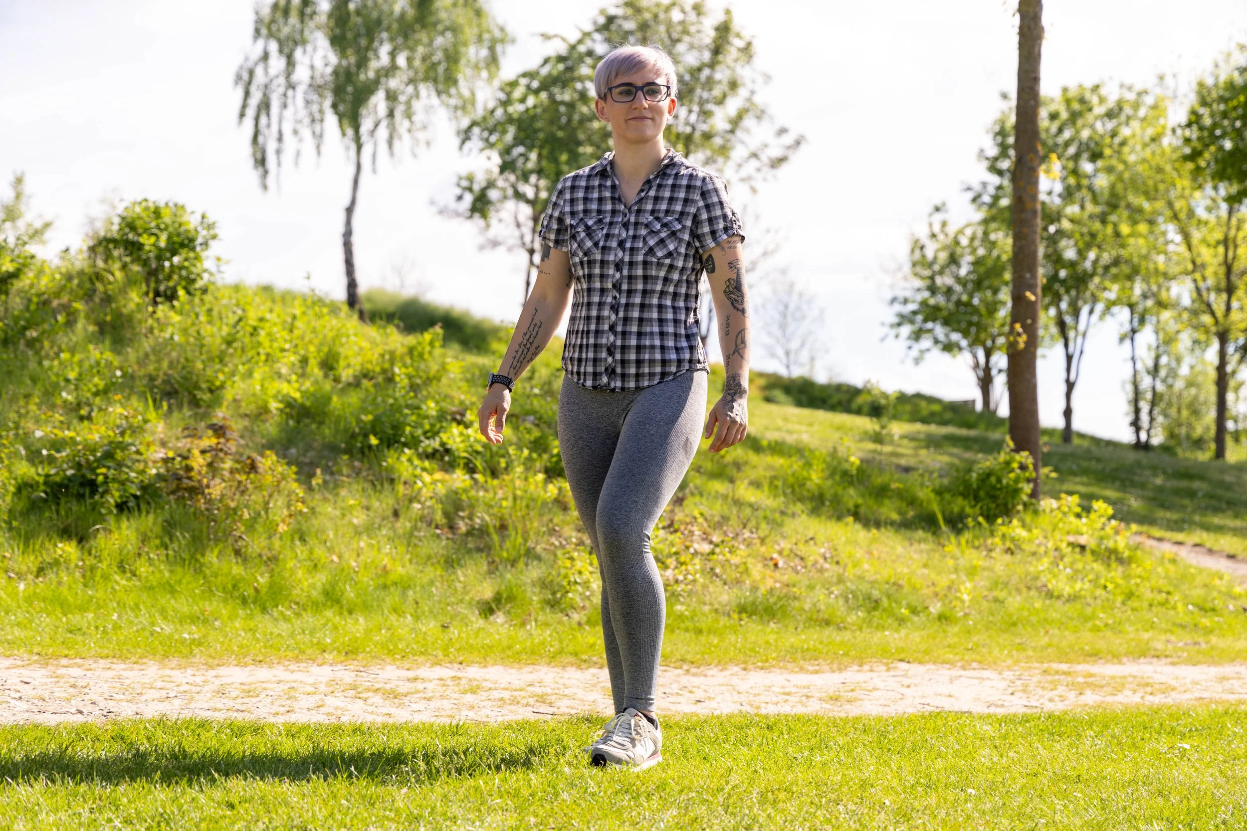Junge Frau mit kurzen, blonden Haaren, Brille und Tattoos, die auf einem Weg in der Natur spaziert.