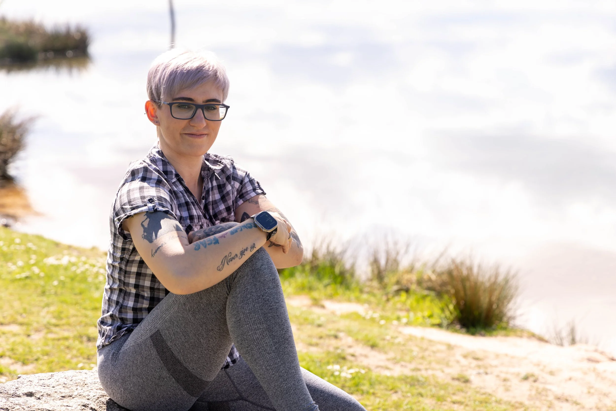Junge Frau mit Kurzhaarfrisur, Brille, Tattoos und Fitness-Tracker sitzt im Park bei Sonnenlicht.