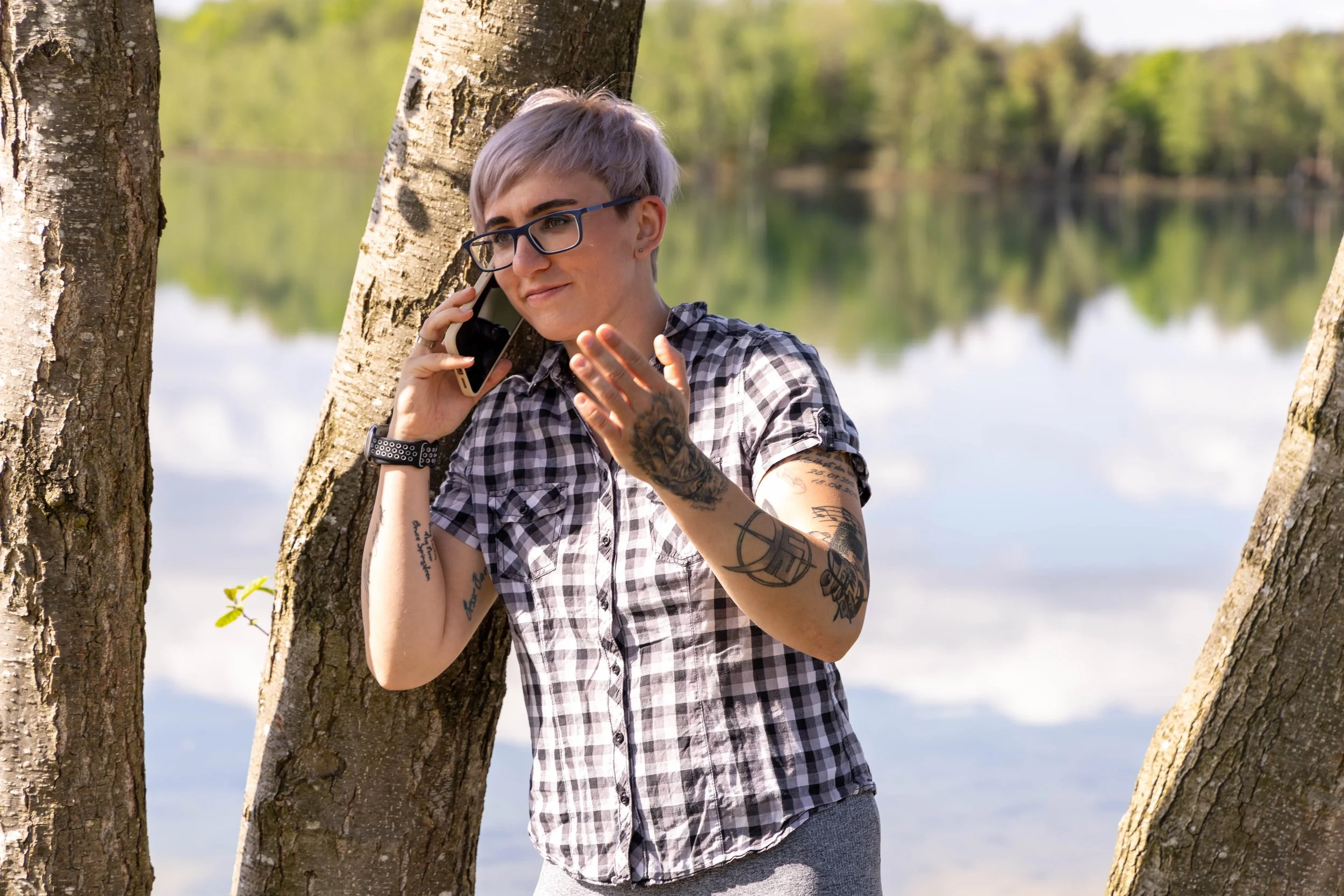 Junge Person mit kurzen, lilafarbenen Haaren, Brille, Tattoos und kariertem Hemd spricht am Handy, steht zwischen Bäumen an einem Fluss oder See, bei Sonnenschein.