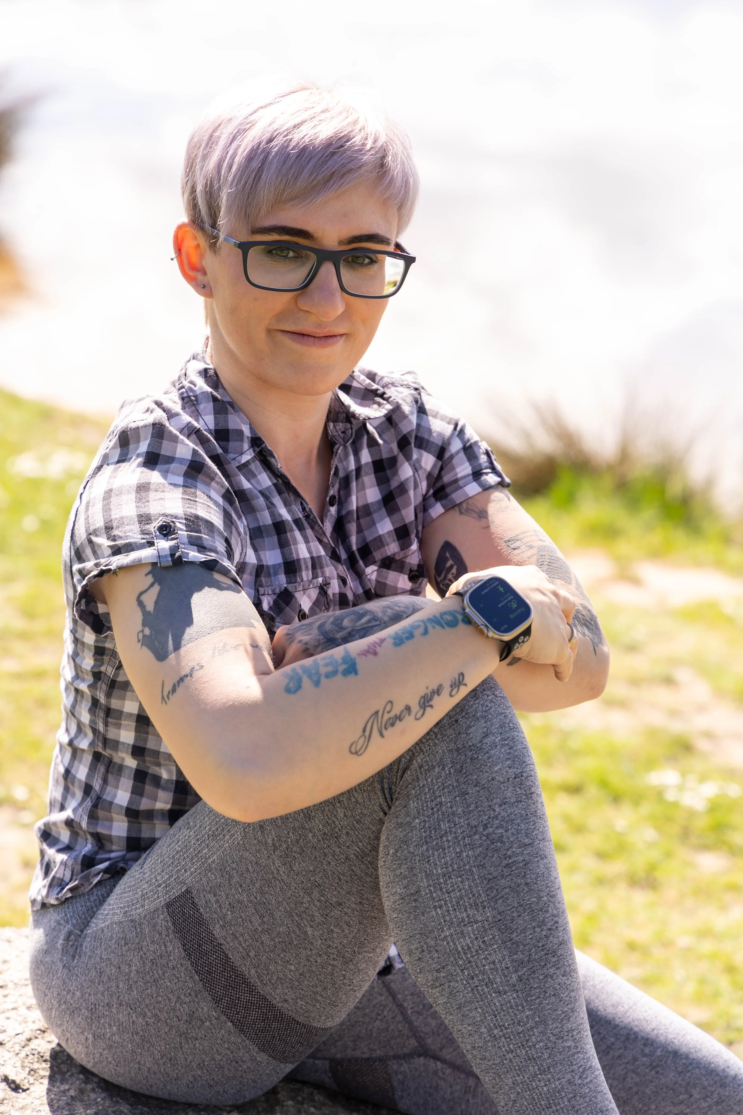 Junge Frau mit Kurzhaar und Brille trägt Sportuniform und sitzt im Park mit Blick auf eine Smartwatch am Handgelenk, sichtbare Tattoos auf den Armen, im Freien bei sonnigem Wetter.