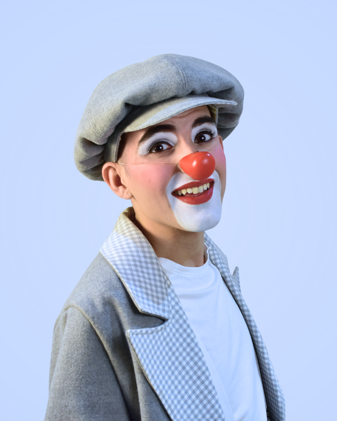 Payaso con maquillaje blanco, nariz roja, gorra gris y chaqueta de cuadros con camiseta blanca, sonriendo.
