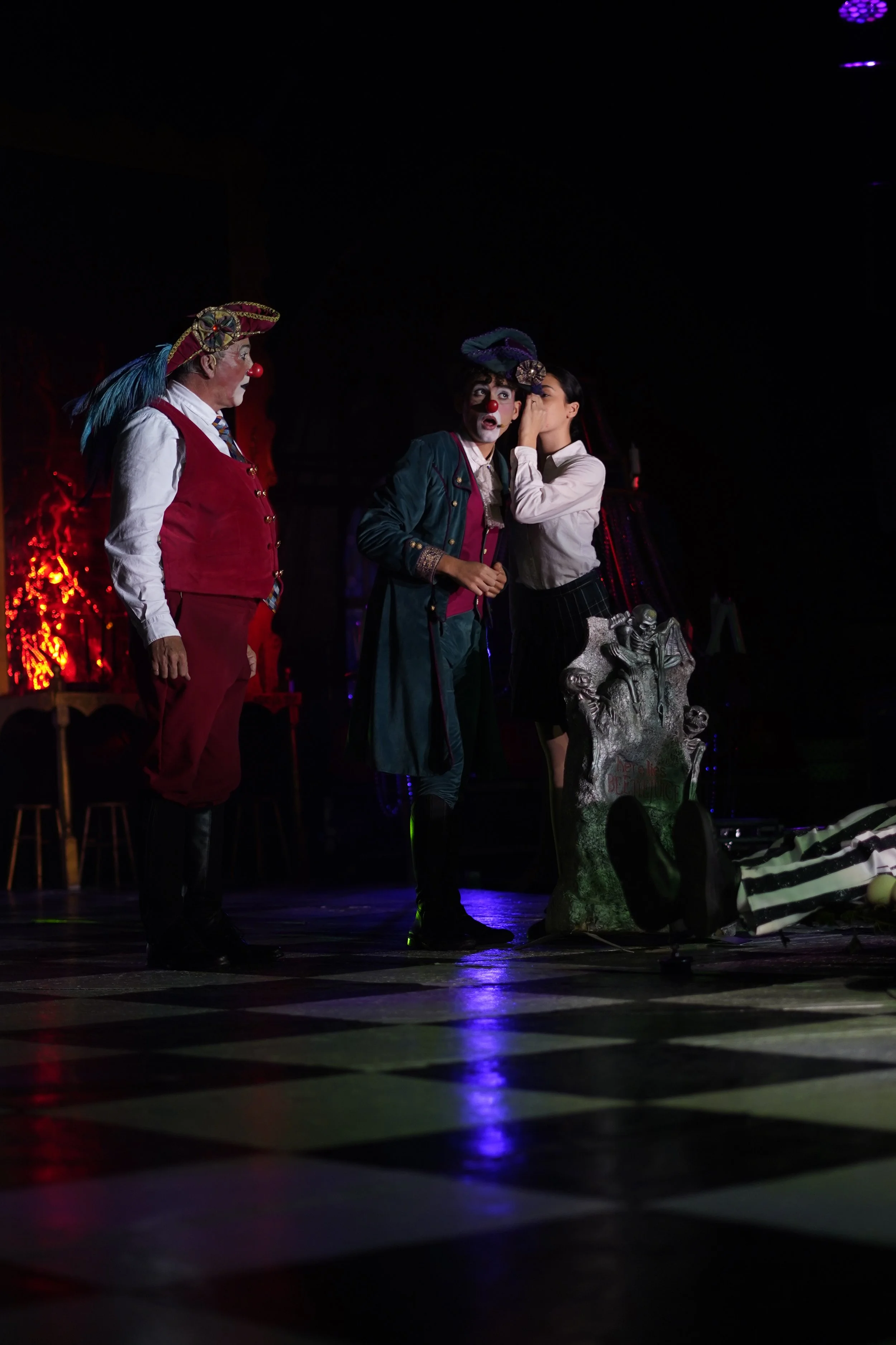 Tres actores en una escena de teatro, uno vestido como payaso y los otros dos en disfraces de payasos, con un fondo oscuro y luces de colores, en un escenario.