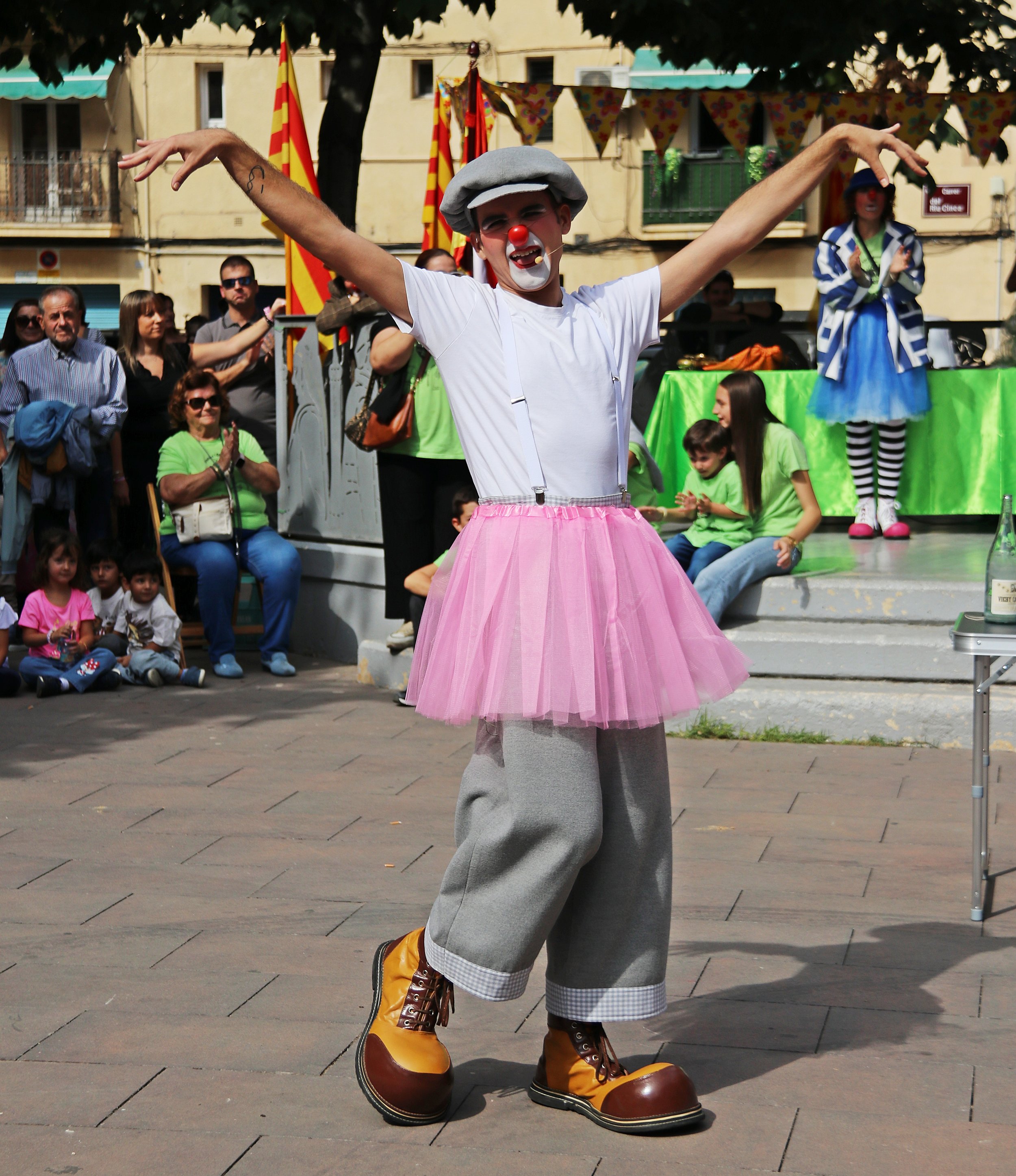 Payaso vestido con camiseta blanca, falda rosa de tul, pantalones gris, botas grandes amarillas y gris, con maquillaje de payaso, nariz roja y expresión divertida, actuando en un evento callejero con público infantil y decoraciones festivas en el fon