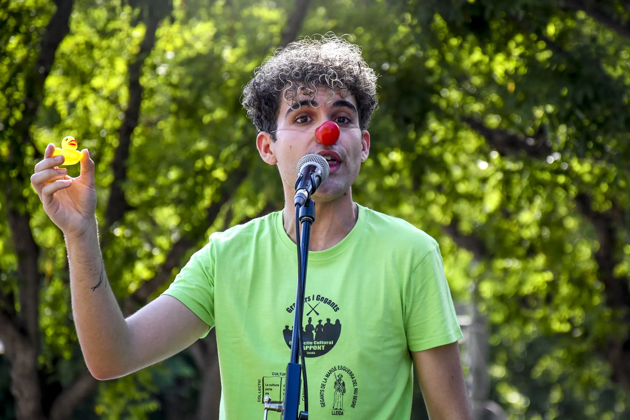 Un hombre con peluca rizada, nariz de payaso roja, camiseta verde y sosteniendo un pato de goma amarillo, hablando frente a un micrófono en un entorno al aire libre con árboles de fondo.