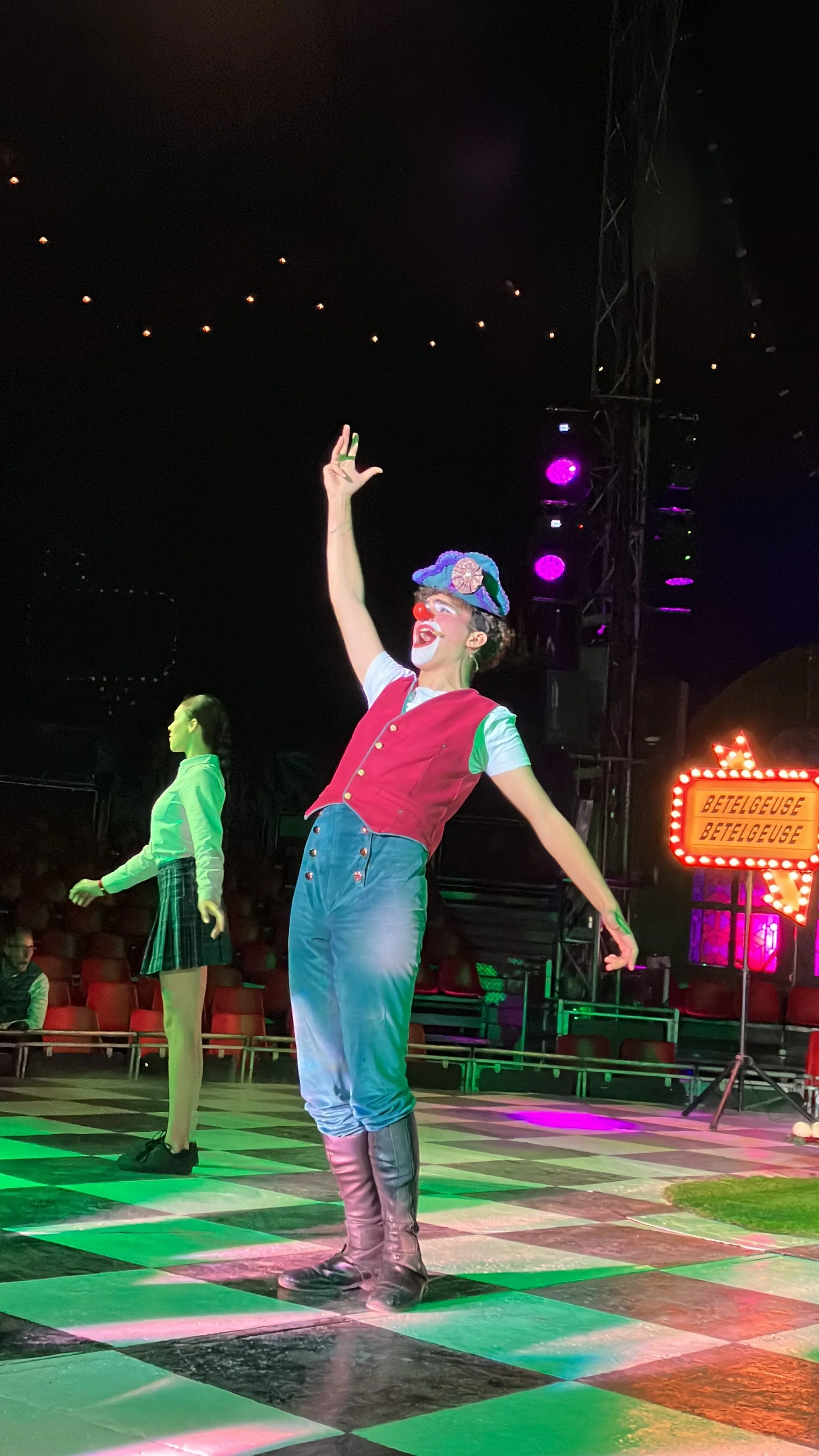 Un payaso vestido con ropa colorida actuando en un escenario, con luces de colores y un letrero que dice 'Betelgeuse'. El payaso tiene maquillaje en la cara, un sombrero decorado y botas grandes. También hay una joven en el fondo vestida con falda y 